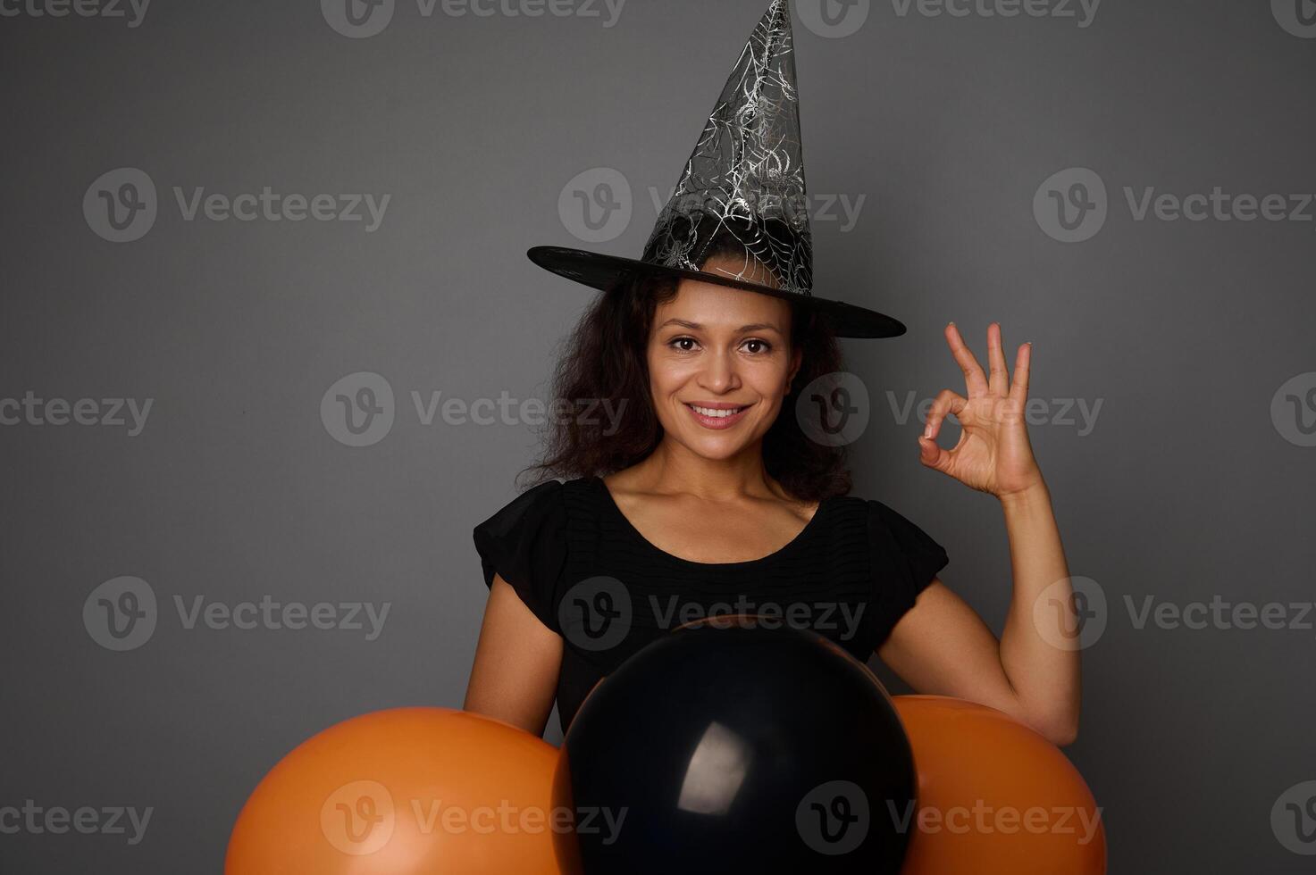 Beautiful smiling Hispanic woman in wizard hat, dressed in witch carnival costume for Halloween party, shows OK sign, poses against gray background with black balloons, copy space photo