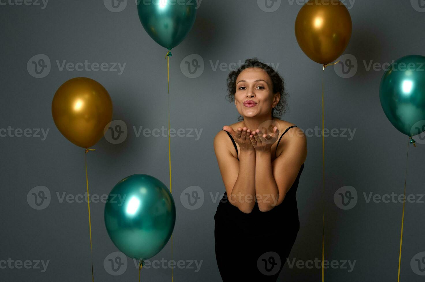 Attractive sensual beautiful woman in black evening dress sends air kisses against gray wall background with golden and green metallic air balloons. Anniversary, Christmas, events concept, copy space photo