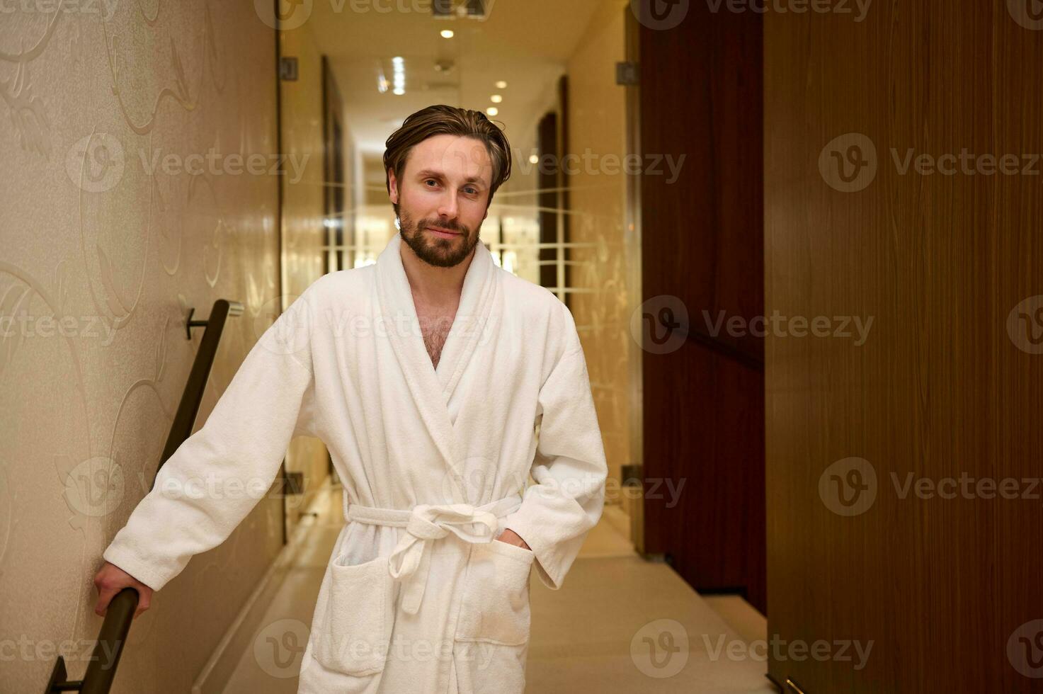 Portrait of a happy relaxed handsome middle aged European man dressed in white terry bathrobe standing on the hallway of a luxury wellness spa resort, smiles looking at camera photo