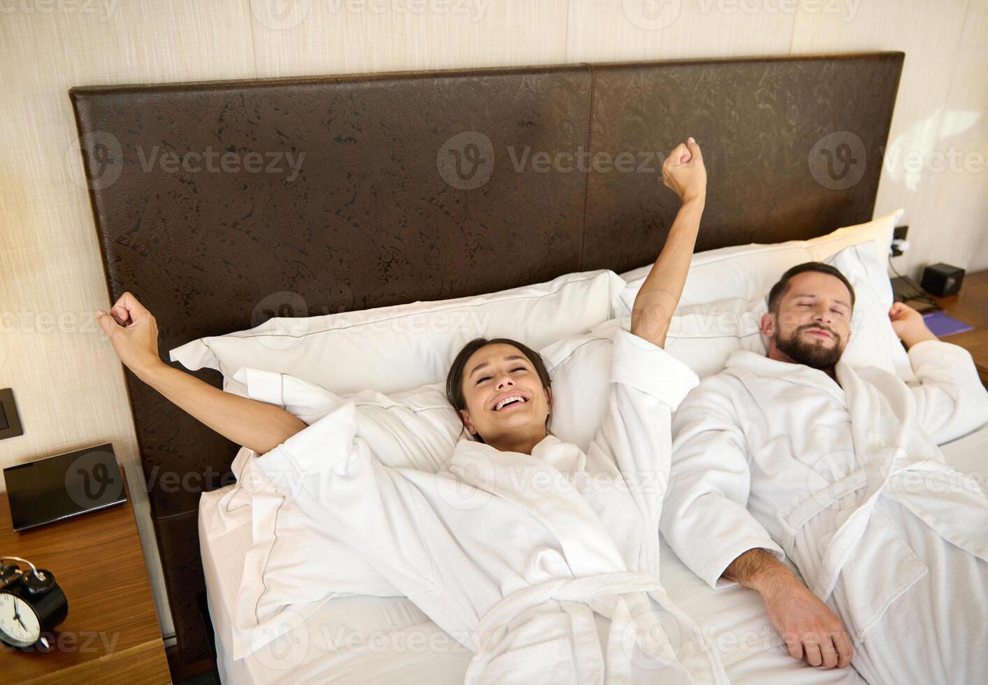 Happy married couple, newlyweds, gorgeous wife and loving husband, lovers stretching in the bedroom after waking up, touching each other's hands and smiling, enjoying the privacy on their honeymoon photo