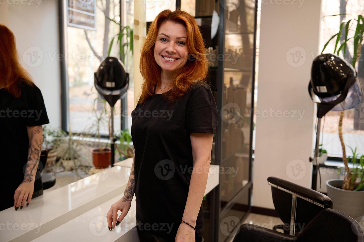 Redhead attractive female hairdresser stylist in black uniform stands near the mirror in a beauty salon, smiling cute toothy smile, looking at the camera while waiting for the client. photo