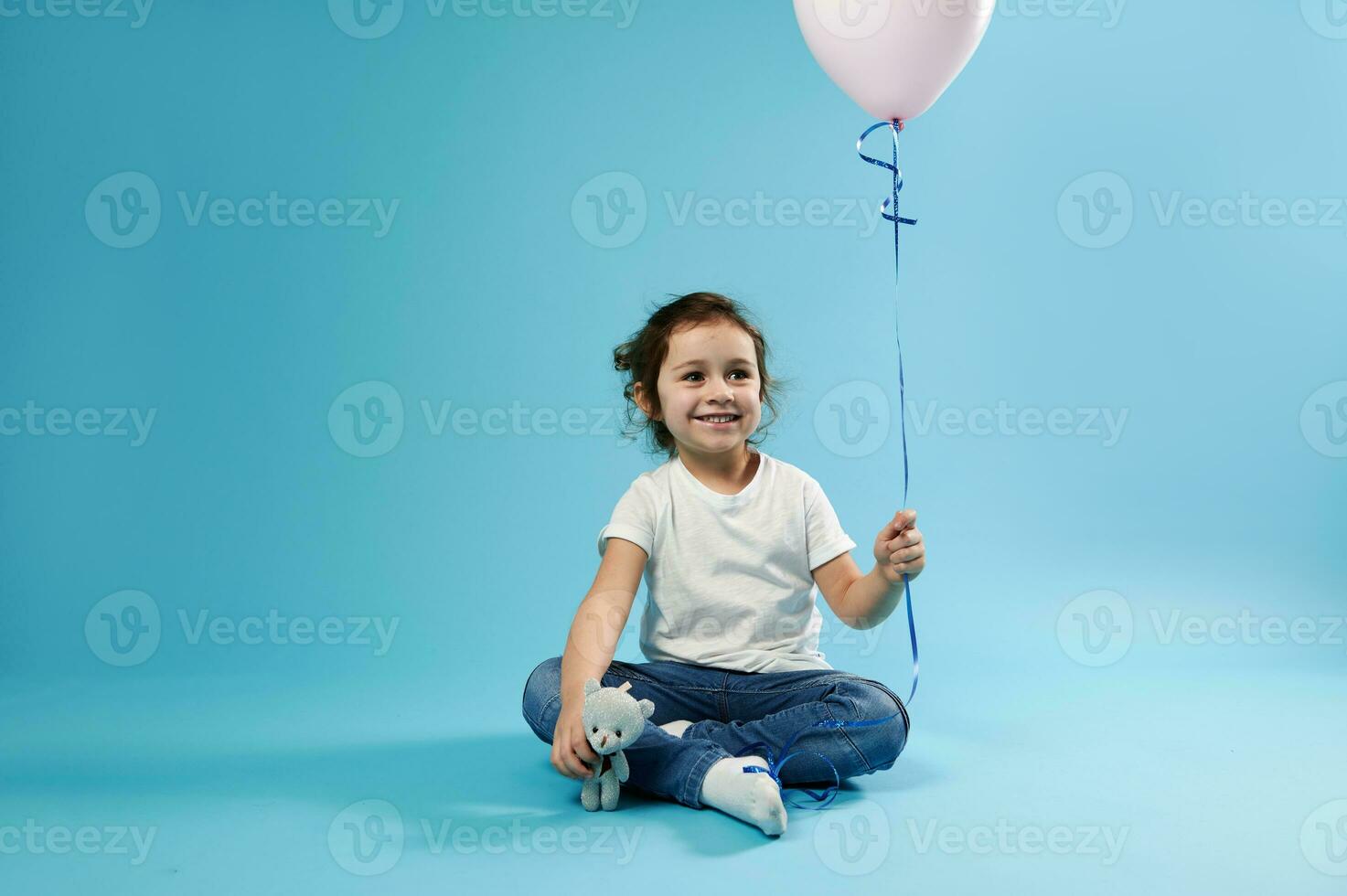 un pequeño linda niña se sienta en un azul antecedentes y sostiene un rosado globo en uno mano y un osito de peluche oso suave juguete en el otro mano foto