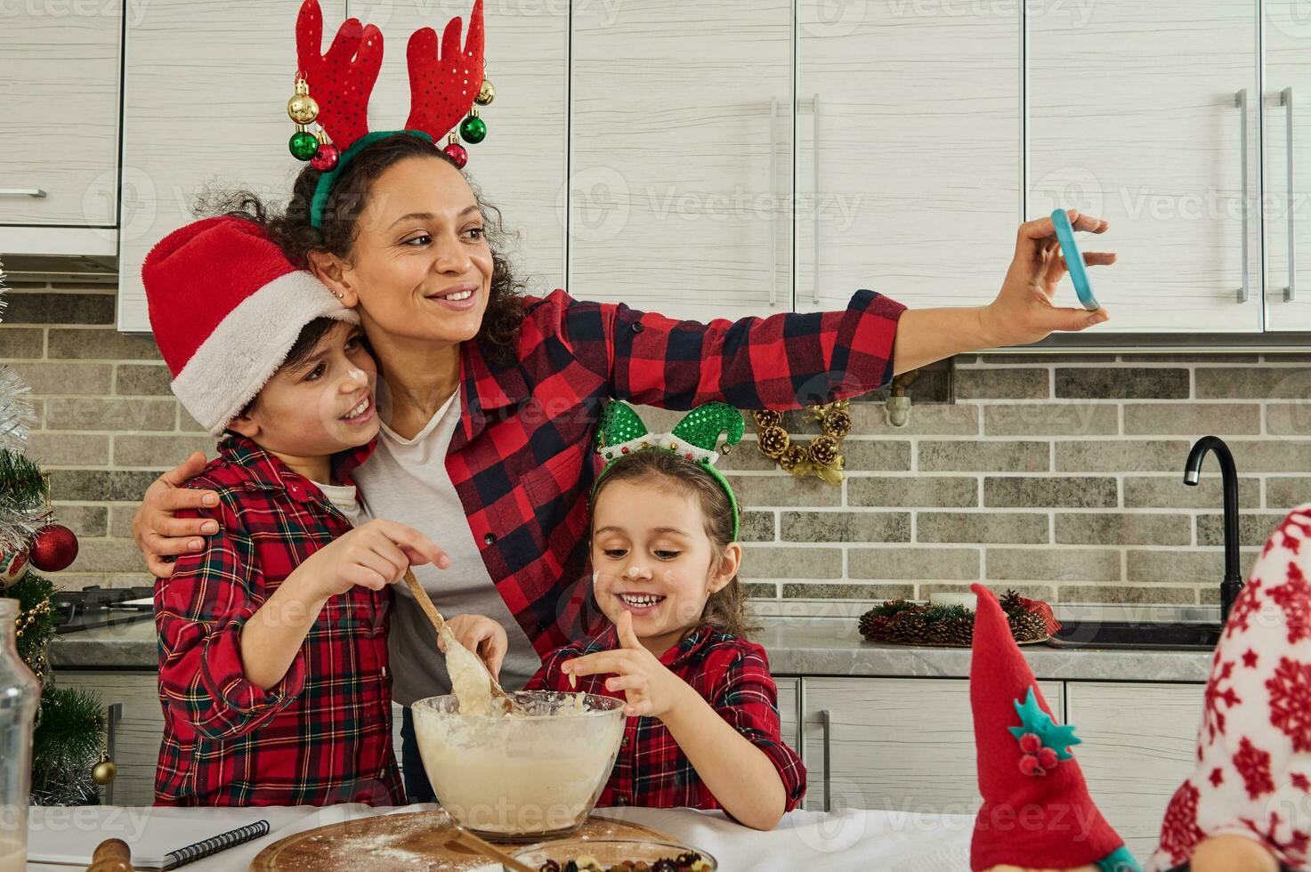 Adorable Caucasian children kneading dough, helping their mother in cooking Christmas bread and cookies. Delighted mom recording video and making selfie, hugging her son and daughter. Happy family photo