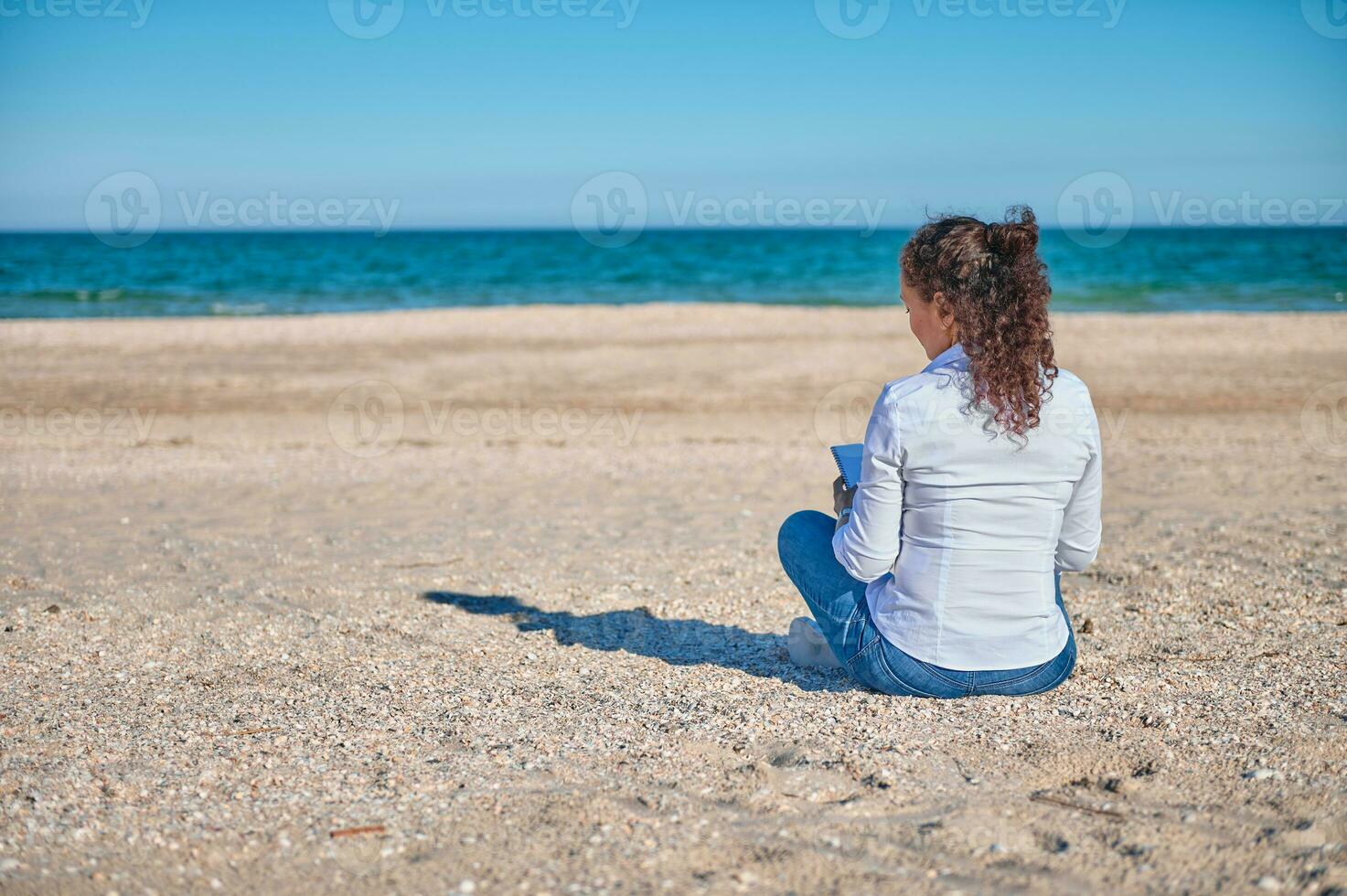 joven morena se sienta en el arena en frente de el hermosa azul mar y sorteos en el playa foto