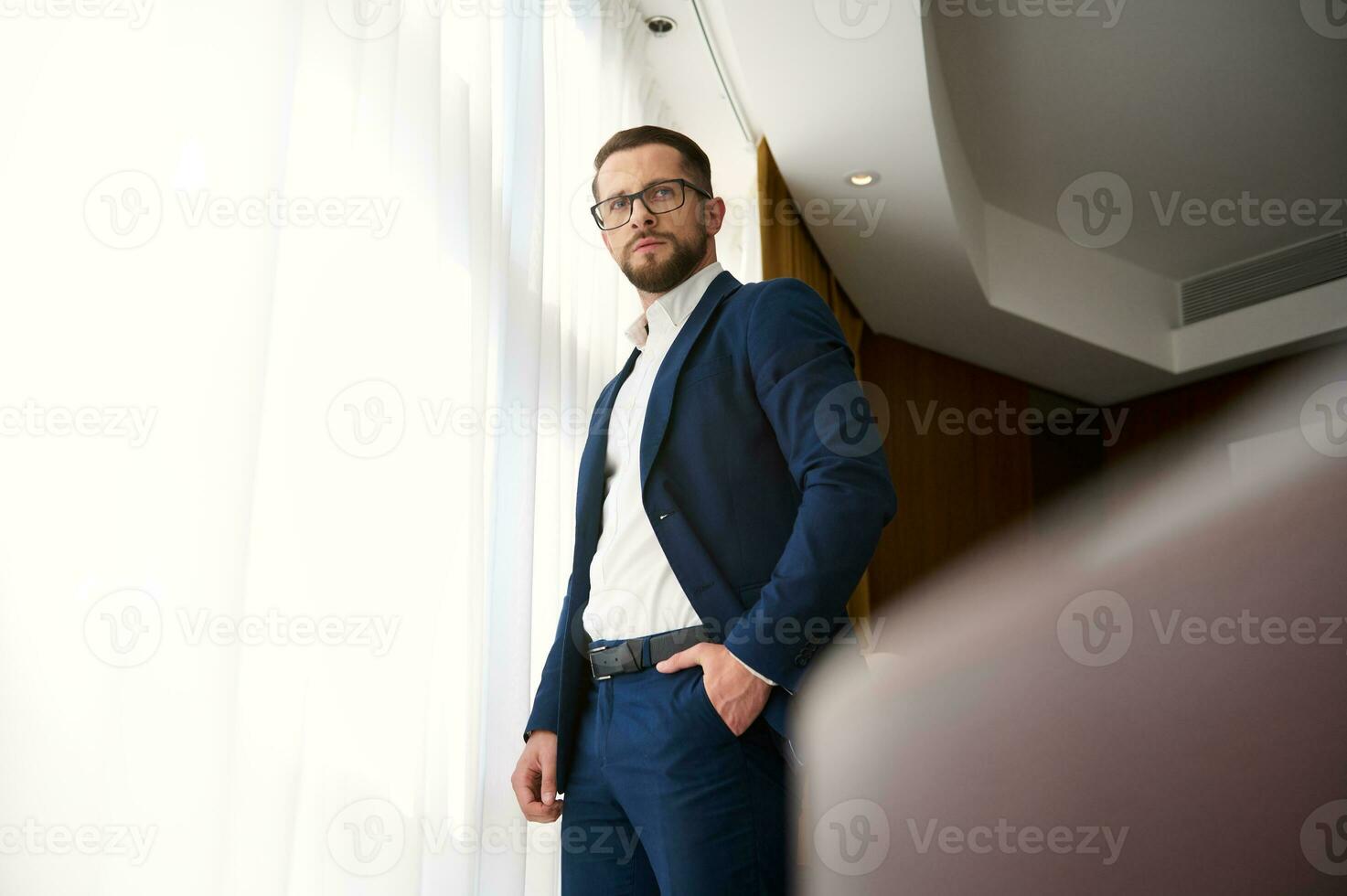 próspero negocio inversor ,elegante exitoso empresario, joven empresario en negocio traje y los anteojos durante un negocio viaje, soportes con manos en bolsillo a el ventana en un hotel habitación foto