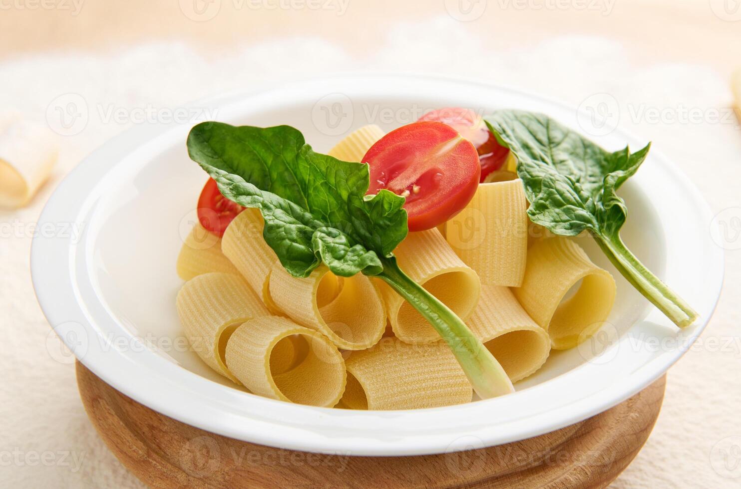 Close-up of traditional Italian whole wheat pasta with halves of tomato and green spinach leaves on a round wooden board on a floured background with copy space photo