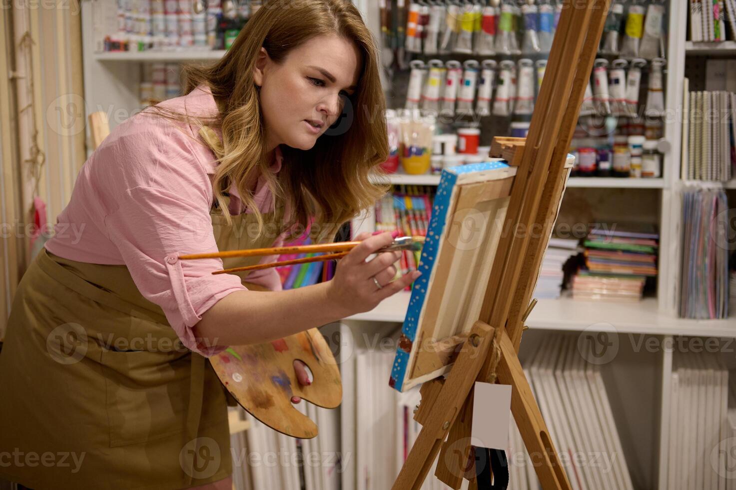 Inspired female painter draws on canvas, standing at easel in a creative art studio against a shelf with oil paint tubes photo