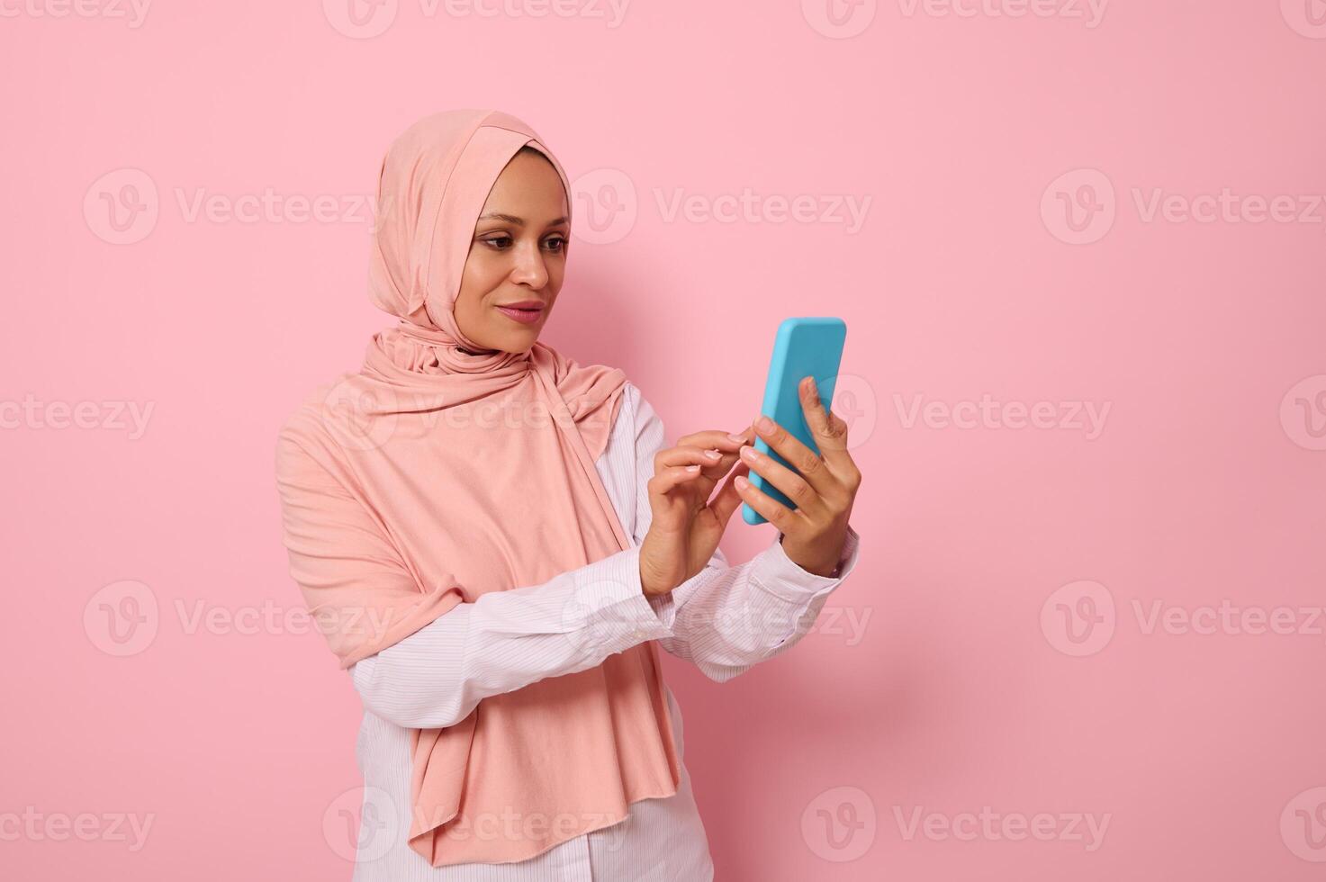 Isolated portrait of confident Arab Muslim middle aged woman in strict religious outfit and covered head in pink hijab texting a message on a mobile phone in her hands, colored background, copy space photo