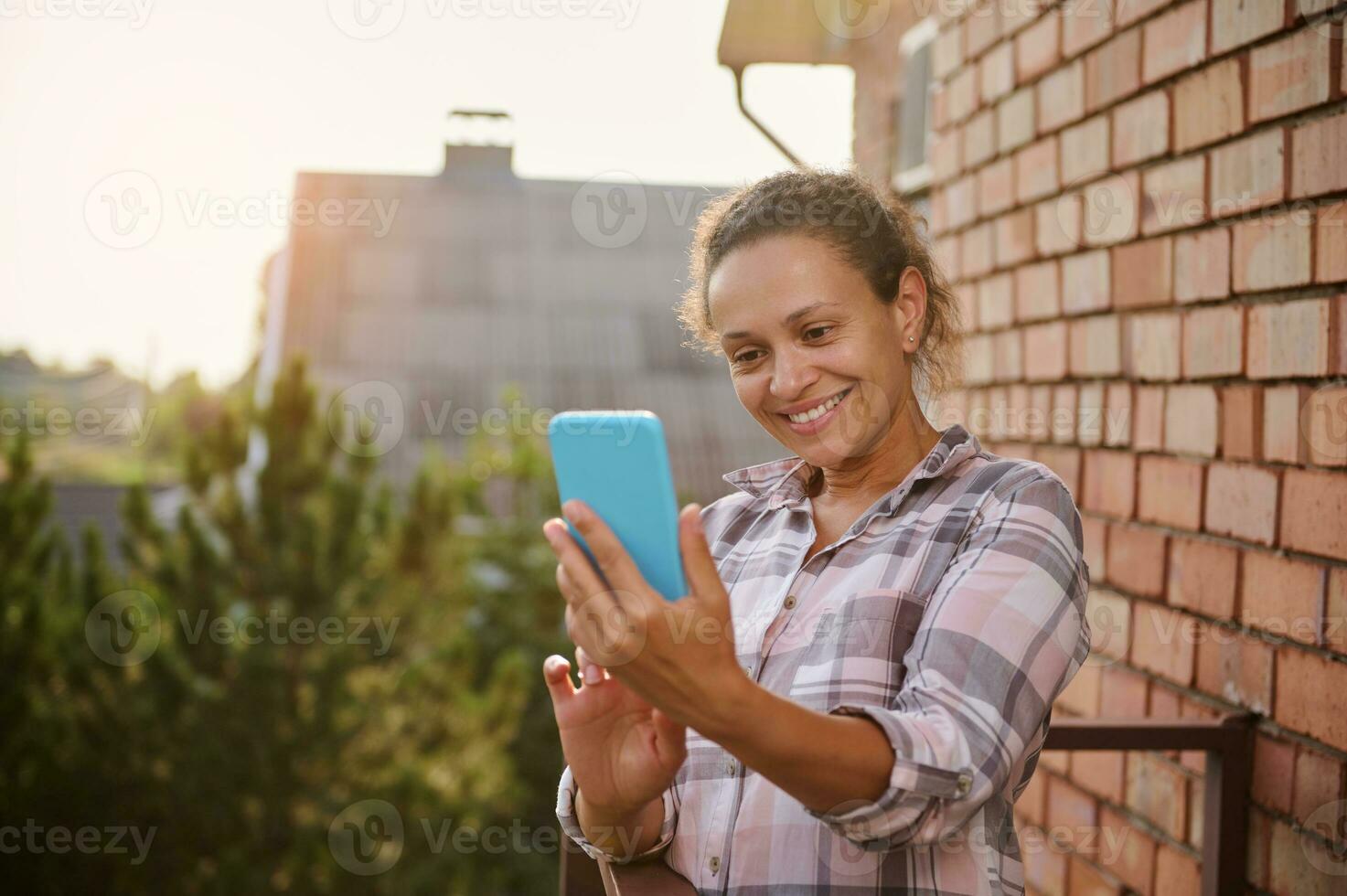 alegre mujer utilizando teléfono inteligente, haciendo selfie en pie en el balcón de un país lado casa en soleado calentar verano día foto