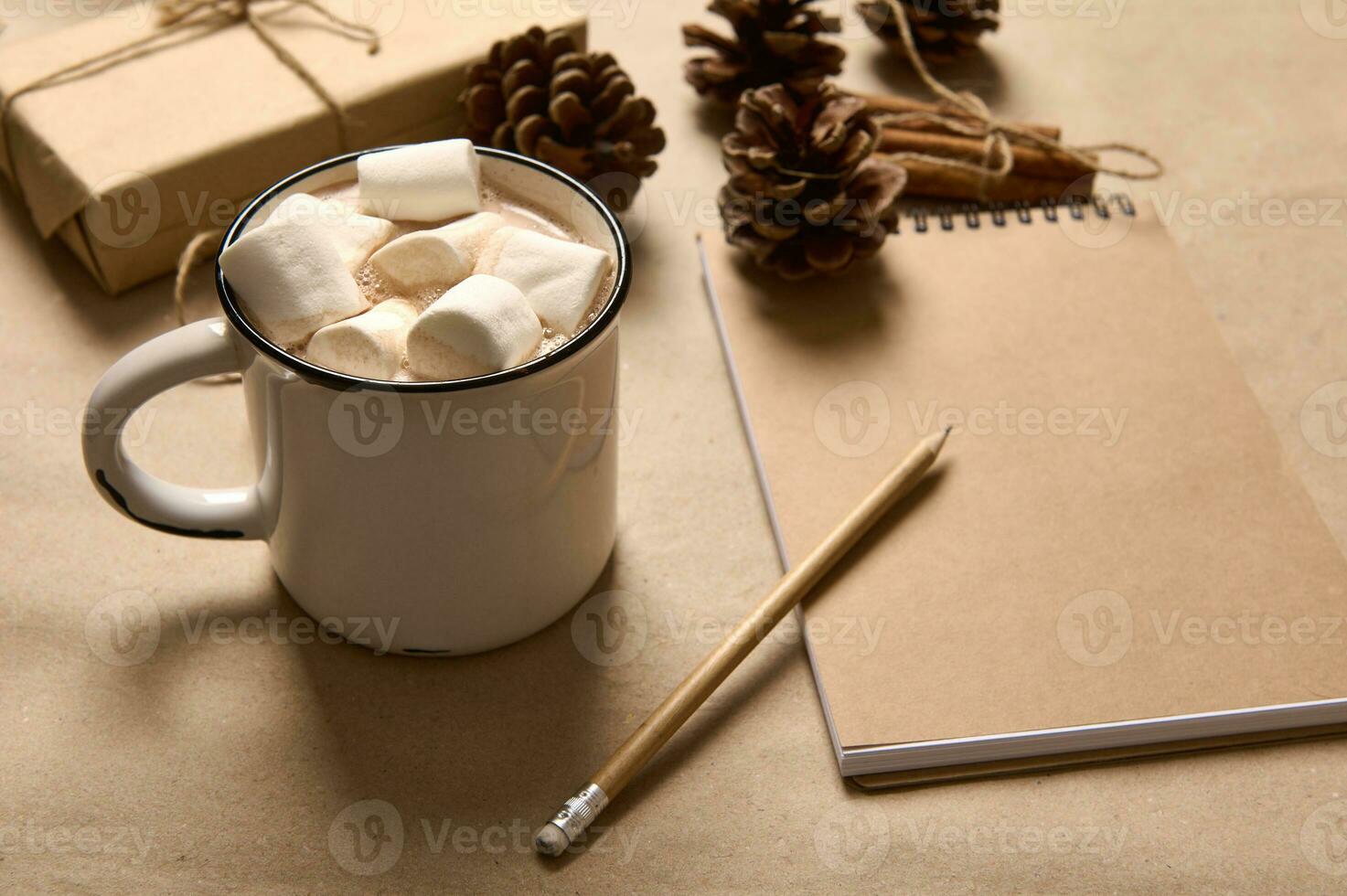 Christmas composition with gift in craft wrapping paper, cinnamon tied with rope, pine cones, wooden pencil on notepad and simple white cup with hot chocolate drink and marshmallows. photo