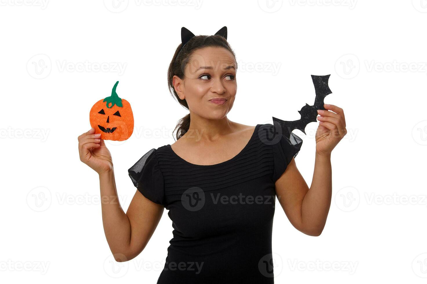 Surprised mysterious smiling Hispanic woman in hoop with cat ears, dressed in black, holds orange handmade felt-cut pumpkin and bat, poses against white background with copy space for Halloween ad photo