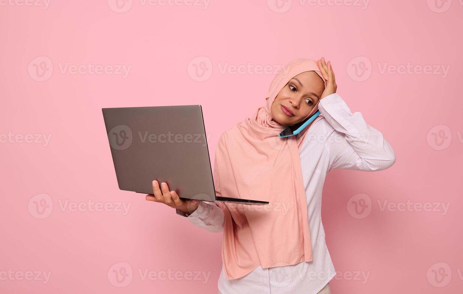Puzzled Muslim Arab woman in pink hijab, with laptop, talking on the phone, holding her head because of multitasking, looking down with sad expression, on colored background with copy space photo