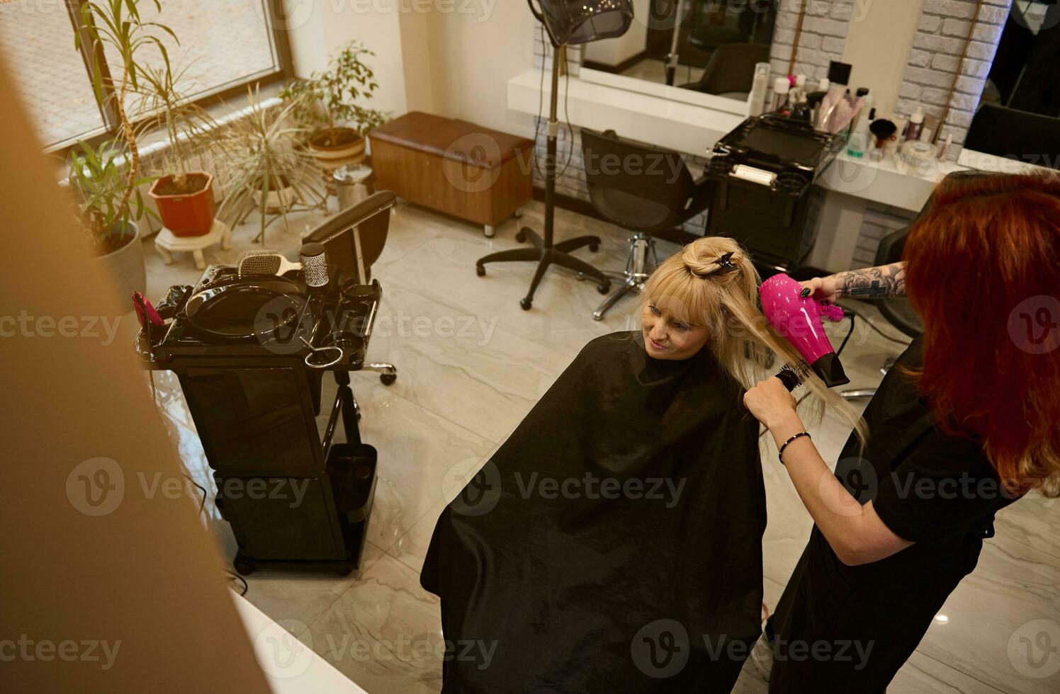 High angle view of redhead female hairdresser combing and drying client hair in beauty parlor, using hairdryer to perform stylish hair style. Fashion, small business and beauty industry concept photo