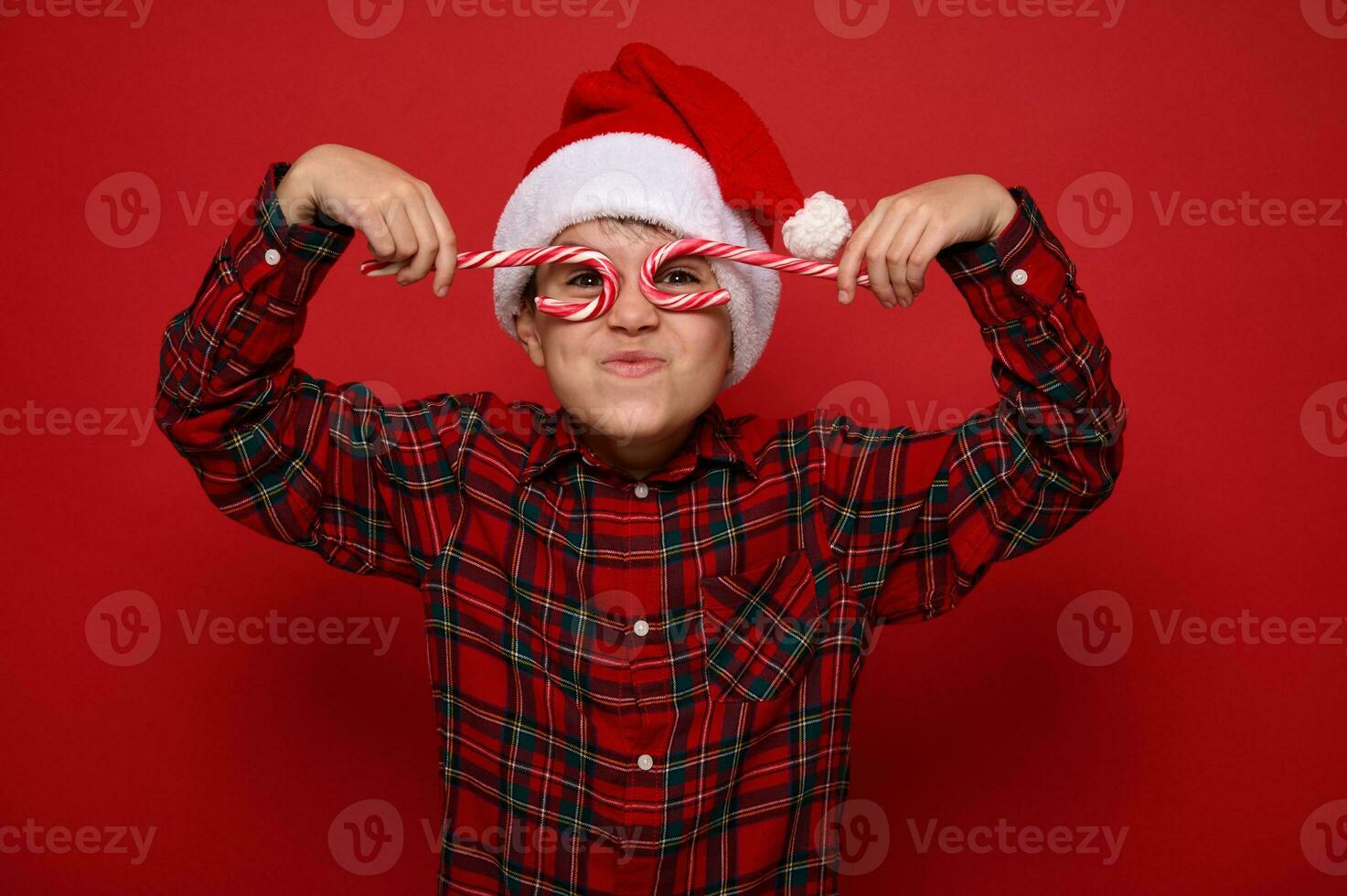 Handsome cute child boy in Santa Claus hat and plaid shirt looks at the camera through Christmas sweet candy canes, holding them imitating eyeglasses, posing on red background with copy space for ad photo