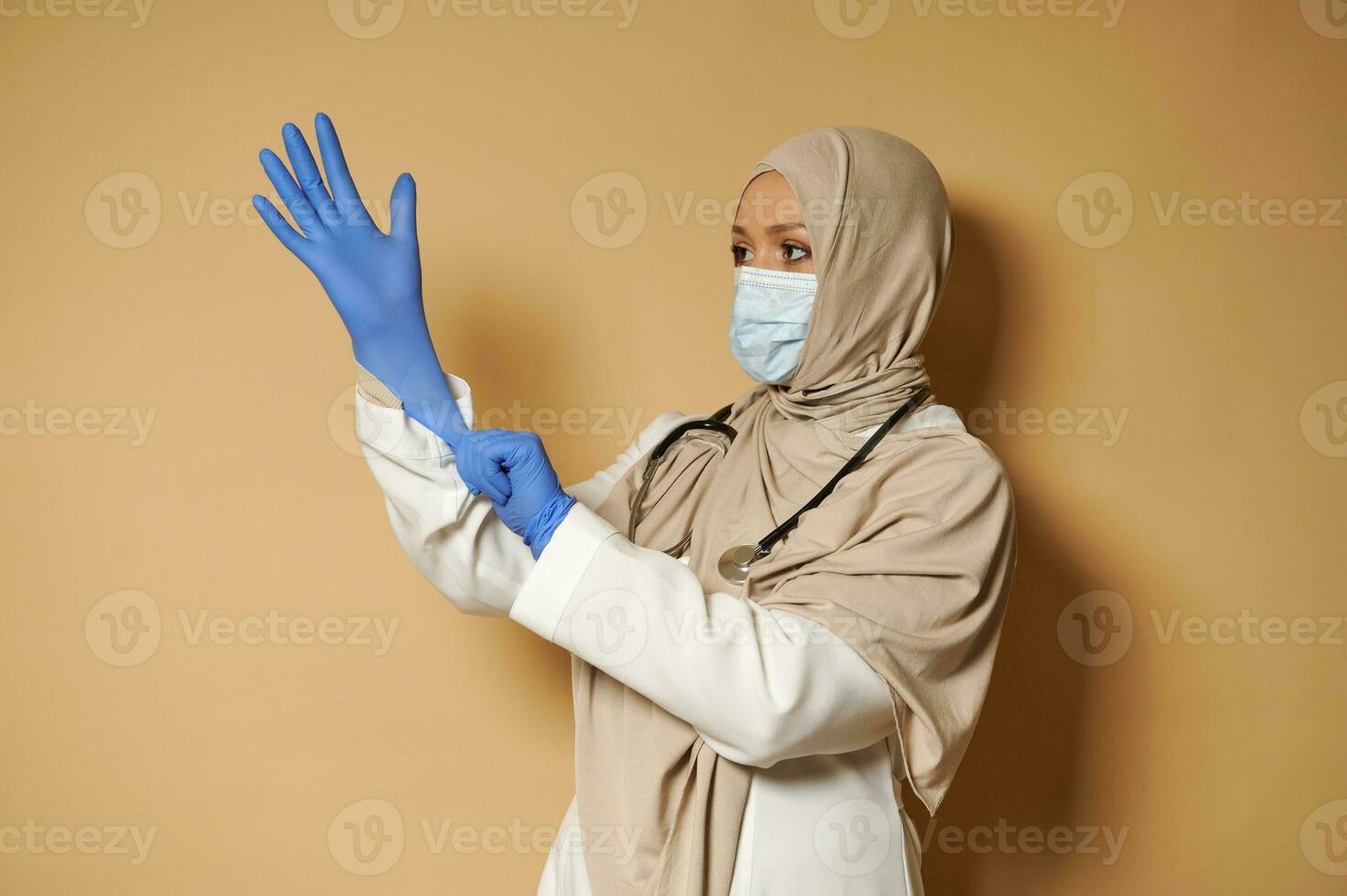 Muslim female doctor in hijab wearing medical protective gloves posing in front of camera on beige background with copy space photo