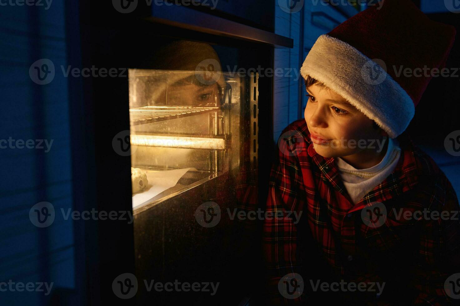 Adorable curious European boy in Santa hat in a red and green plaid shirt sits on the floor and looks through the oven at the traditional German Stollen bread baked on a baking tray photo
