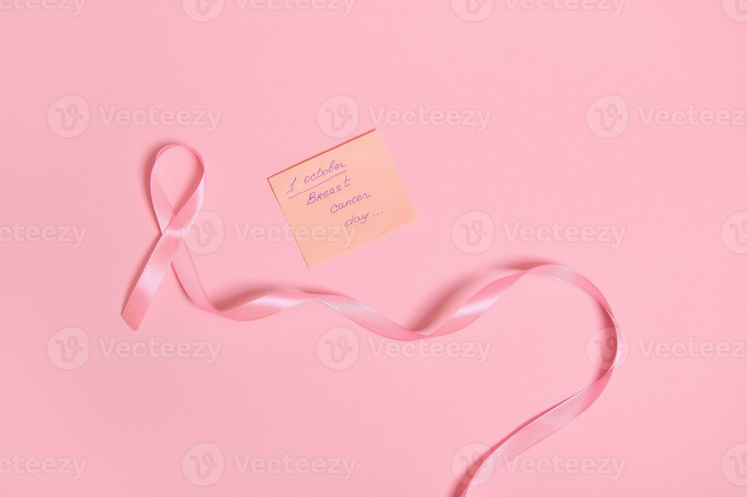 Flat lay of a long pink ribbon with endless one end, and a paper note with lettering 1 October Breast Cancer Awareness Day , isolated on pink background with space for text photo