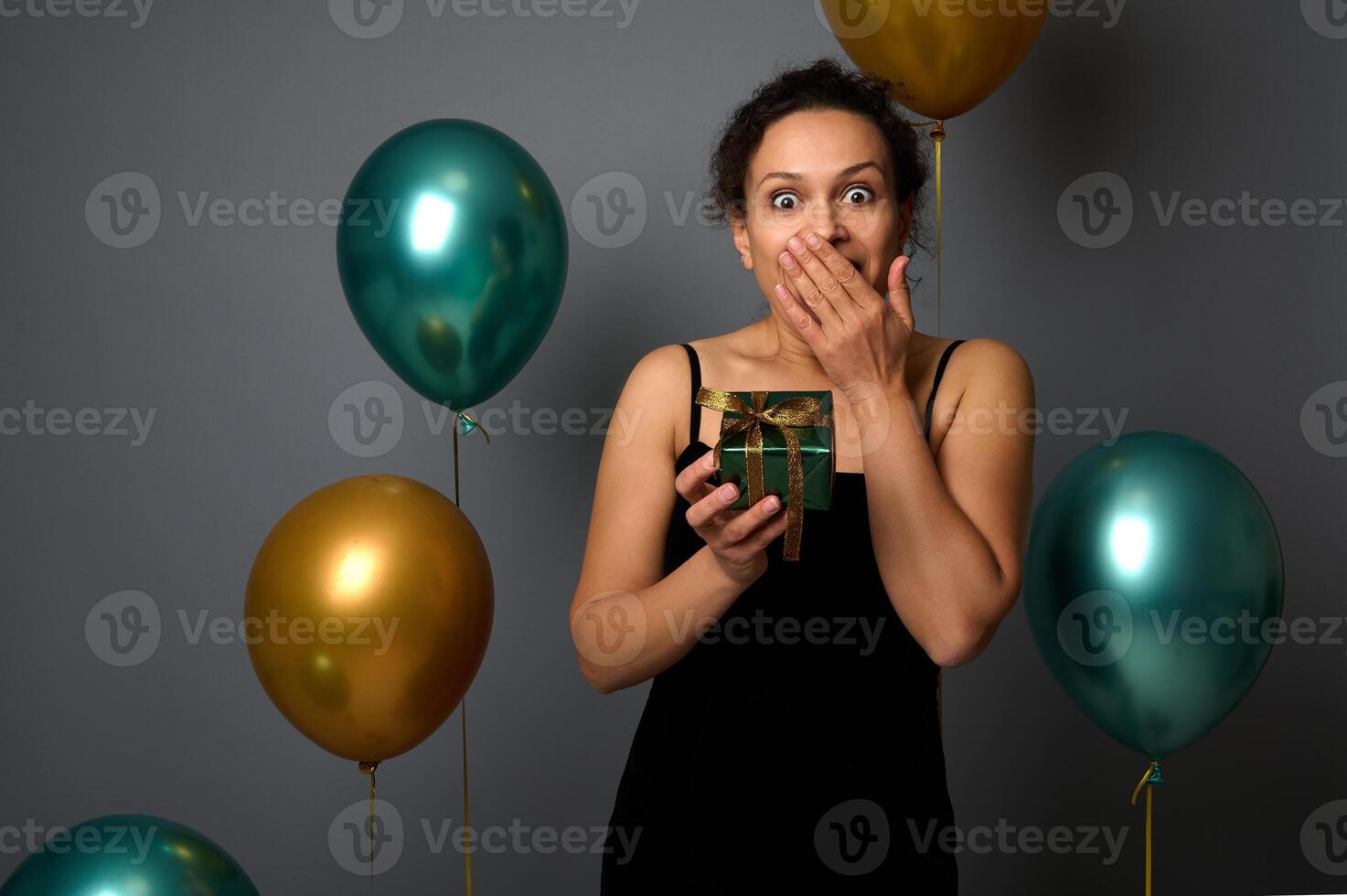 Beautiful woman of African descent, wearing an evening dress, covers her mouth, surprised by a Christmas gift in shiny green wrapping paper an golden bow, isolated over gray background with air balls photo