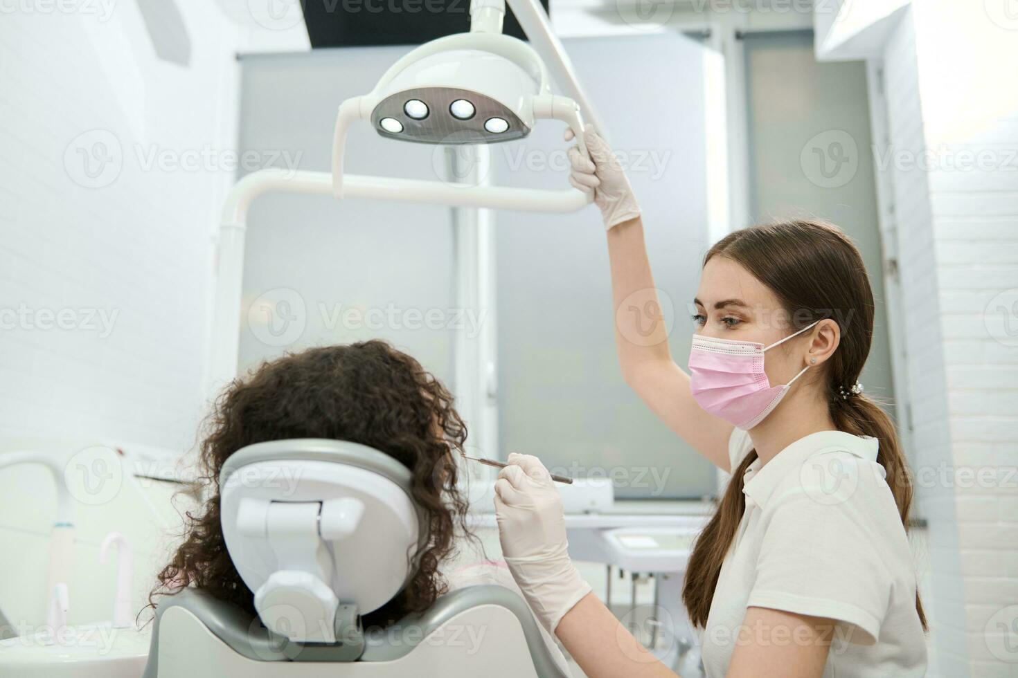 Female dentist in protective medical mask adjusting the lamp before examining the oral cavity of a patient sitting in the dental chair in modern dentistry equipped with modern professional equipment photo