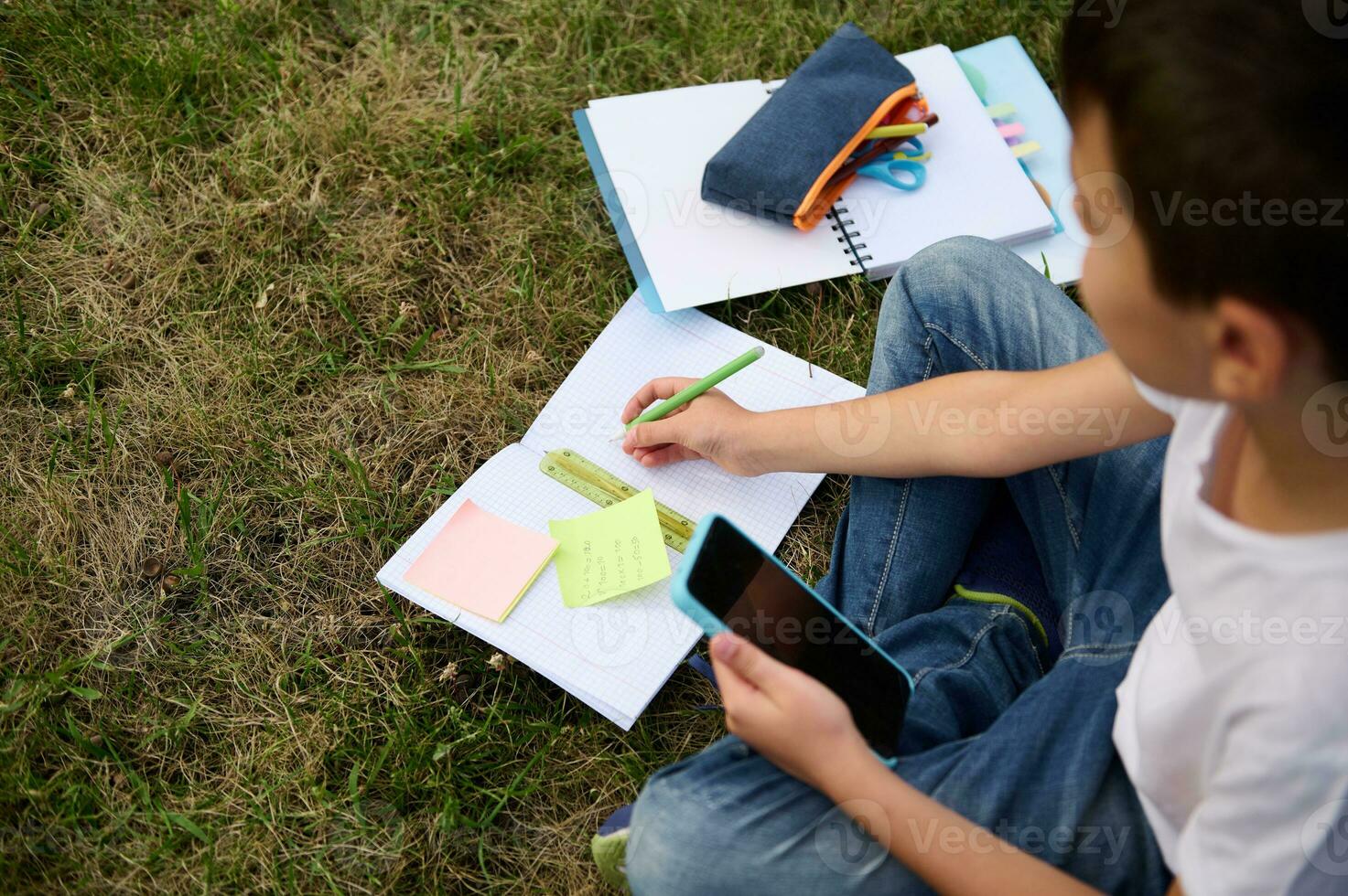 Overhead view of a cute clever school boy studying in the park, sitting on green grass and solving mathematics task, using smartphone and mobile applications, making notes on notebook and workbook. photo