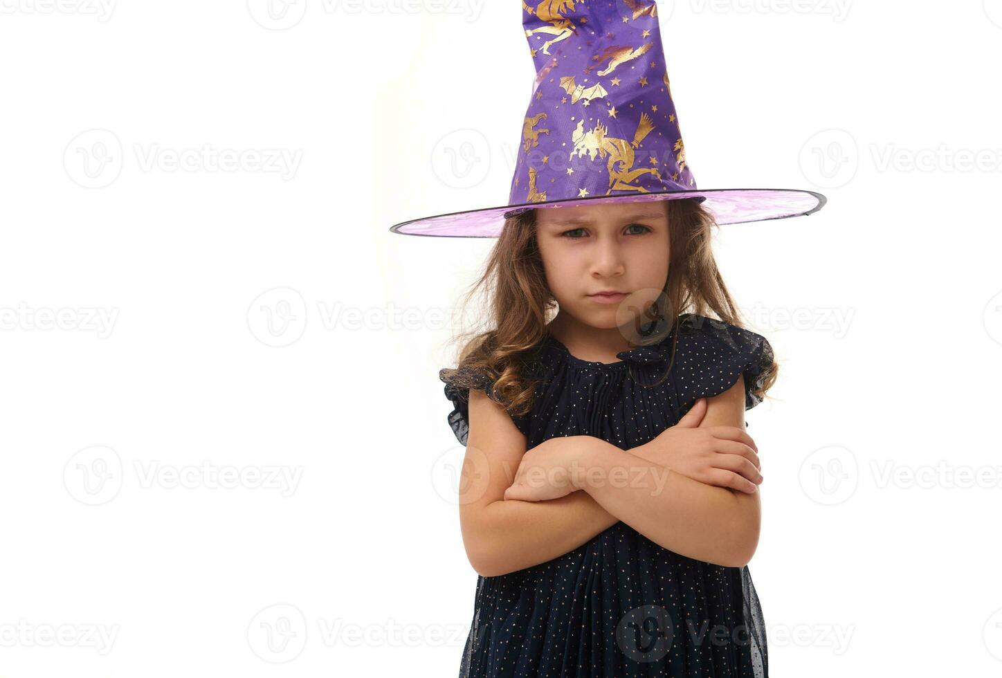 Portrait of pretty little witch angry upset girl wearing a wizard hat and dressed in stylish carnival dress, looking at camera posing with crossed arms against white background, copy space. Halloween photo