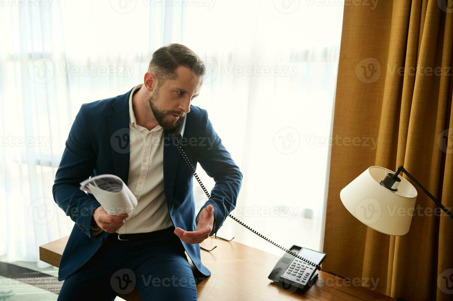 joven hermoso caucásico empresario, discutiendo mientras hablando en teléfono fijo teléfono, participación documentos en su mano y gesticulando con mano durante negociaciones por teléfono, sentado en el escritorio esquina en hotel foto