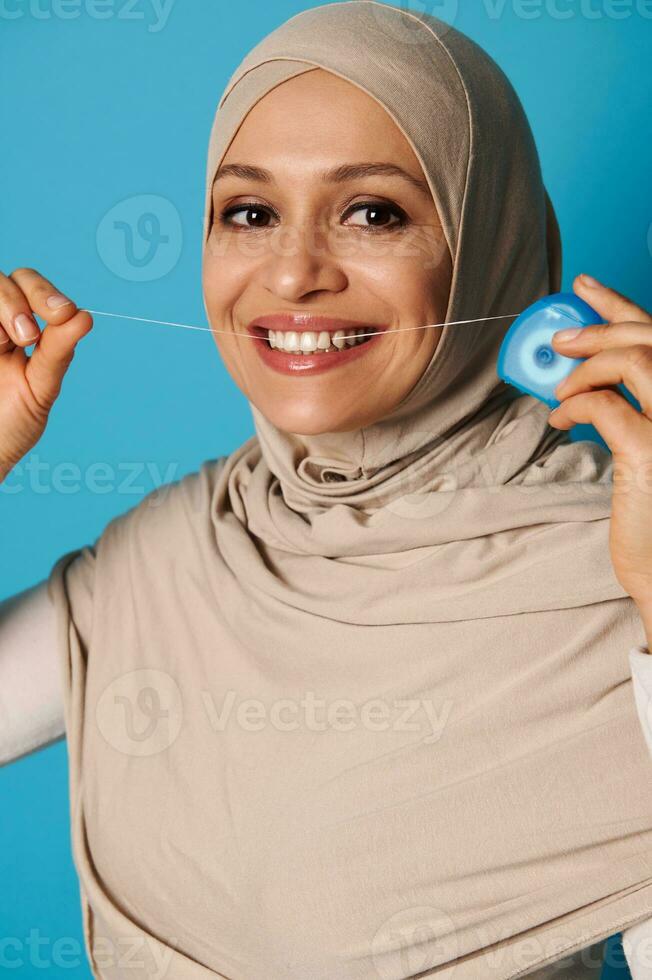 Young Muslim woman with beautiful and healthy smile flossing teeth on color background photo