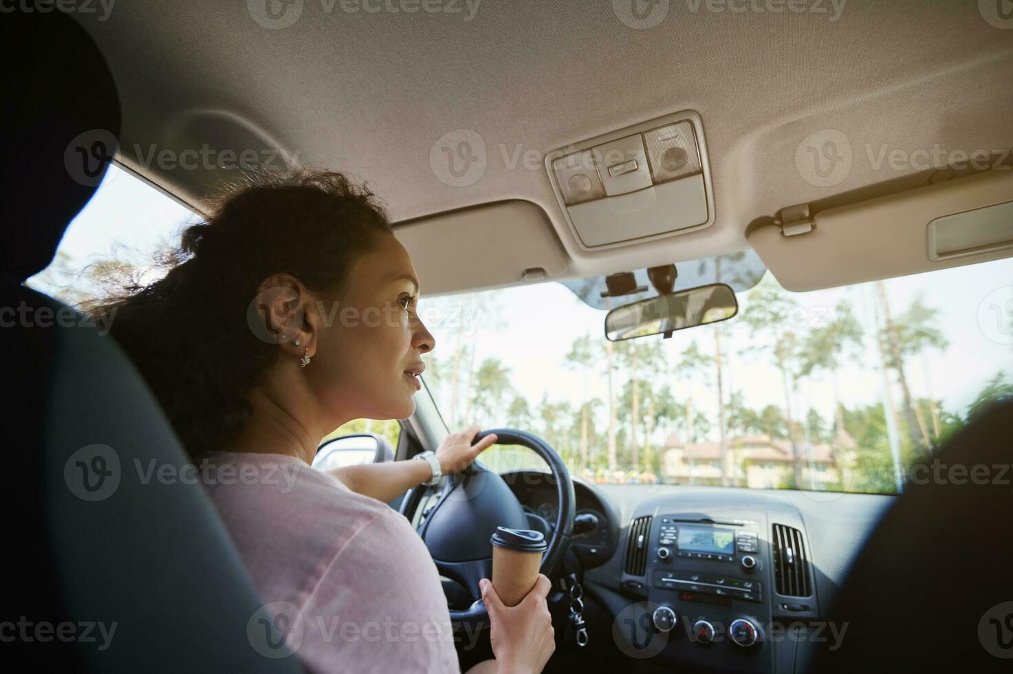 maduro conductor mujer mirando a el lados y poniendo su mano con para llevar papel taza en un direccion rueda en el Moviente coche foto
