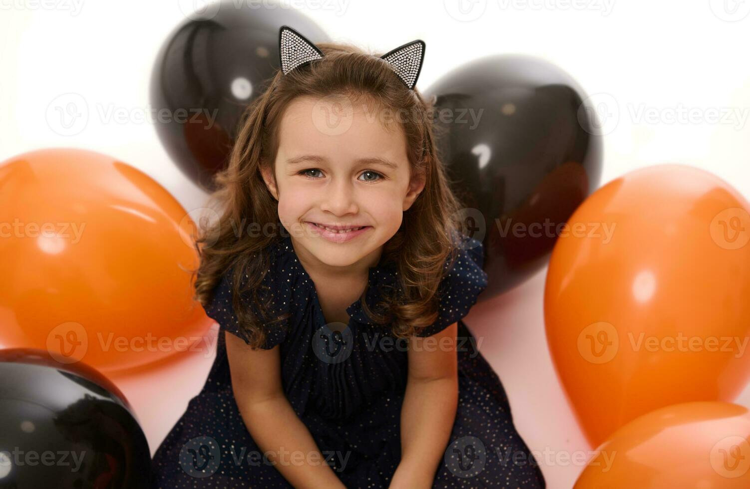 Close-up portrait of a cute little girl smiling with beautiful toothy smile looking at camera on the white background with lying down inflated black and orange colored balloons. Halloween concept photo