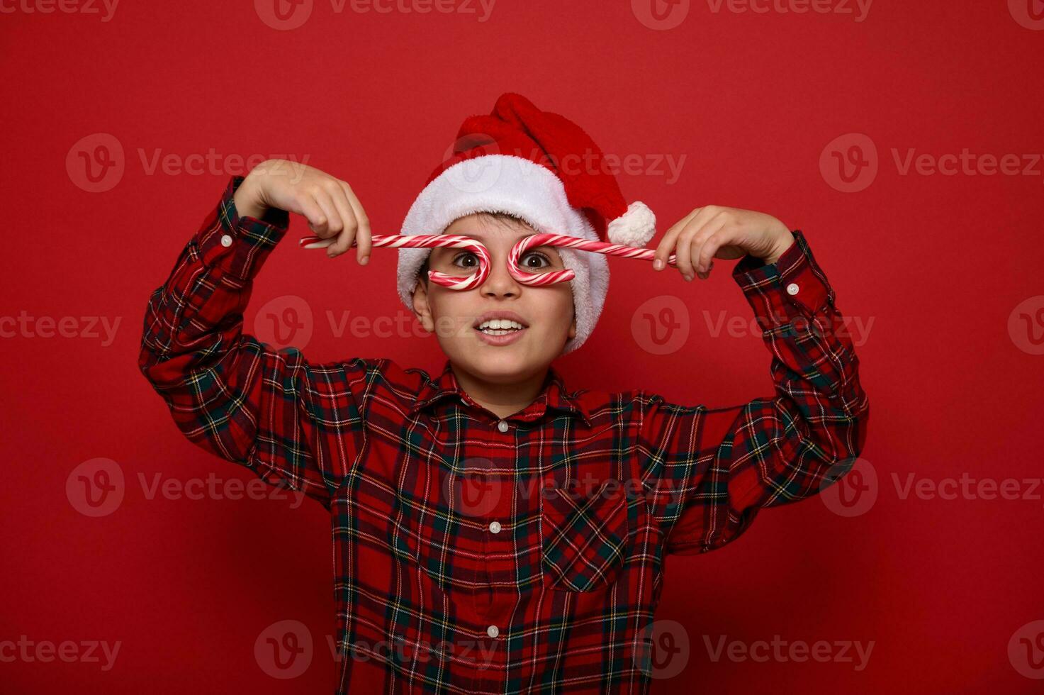 Adorable boy in Santa Claus hat and plaid shirt looks at the camera through sweet lollipops, holding them imitating eyeglasses, posing on a colored background with copy space for Christmas advertising photo