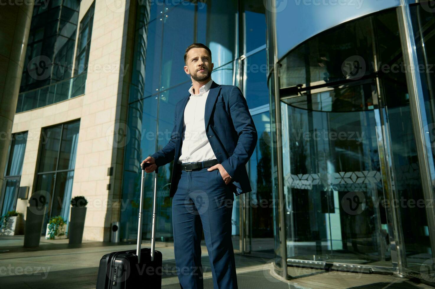 Portrait of a successful young businessman wearing casual business suit stands with a black suitcase at the glass entrance to the hotel, looks confidently to the side, holding his hand in his pocket photo