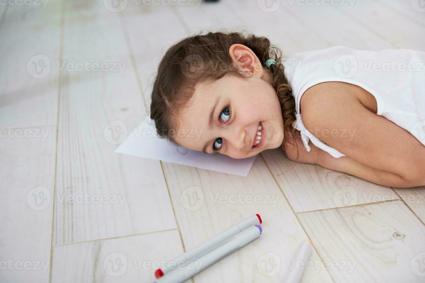 Close-up portrait lovely little girl smiling at camera, lying on the floor while drawing picture with watercolor markers photo