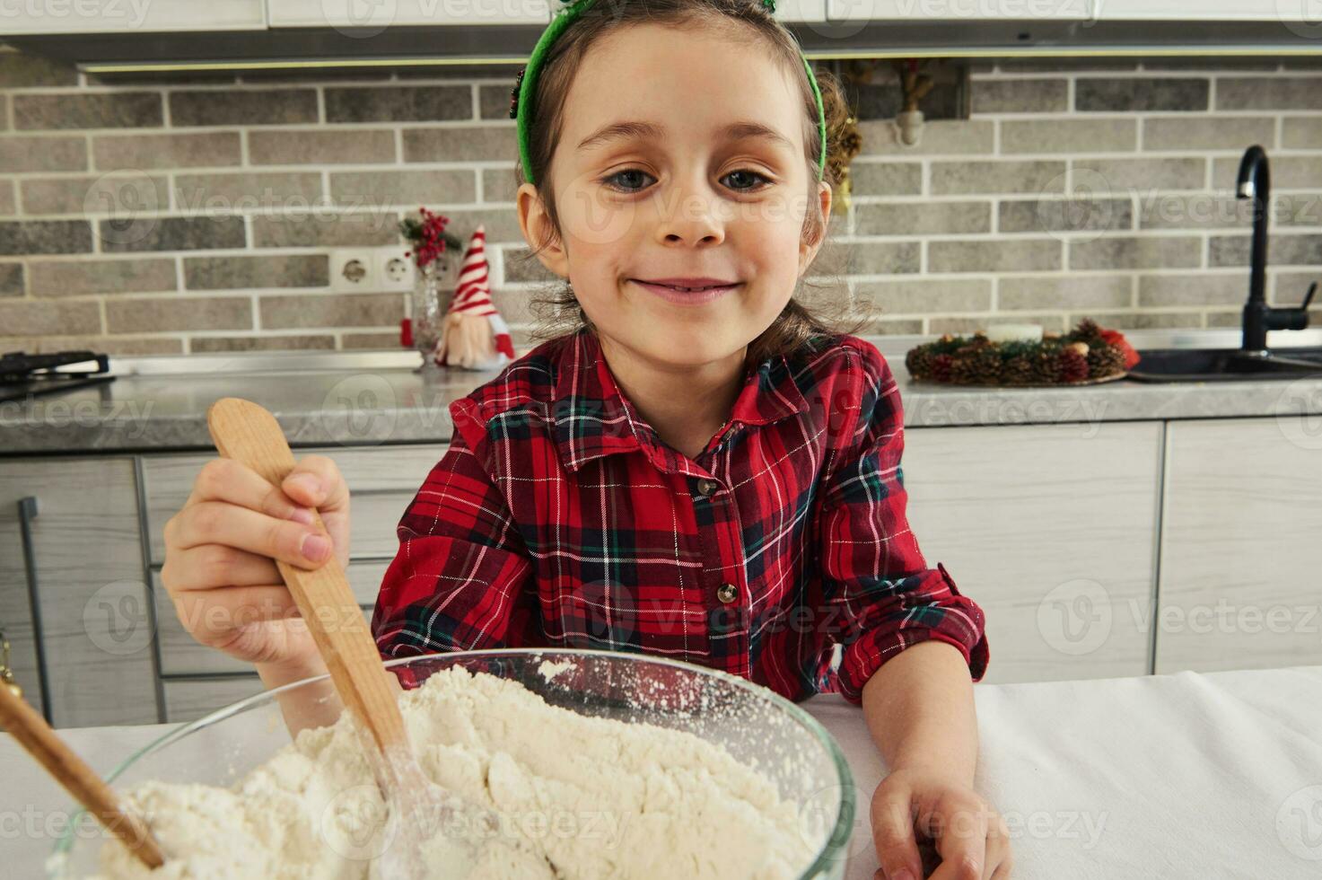 de cerca de un hermosa pequeño niña en rojo y verde a cuadros vestir y duende aro en su cabeza sonriente mirando a cámara, mezcla ingredientes en vaso cuenco con de madera cuchara, Cocinando Navidad Pastelería foto