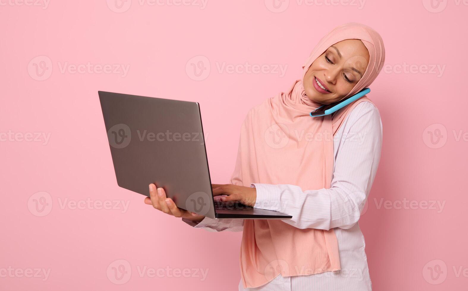 Concept of a busy Arab muslim business woman, freelancer in hijab, talking on a mobile phone while working on a laptop at the same time. Isolated on pink background with copy space photo