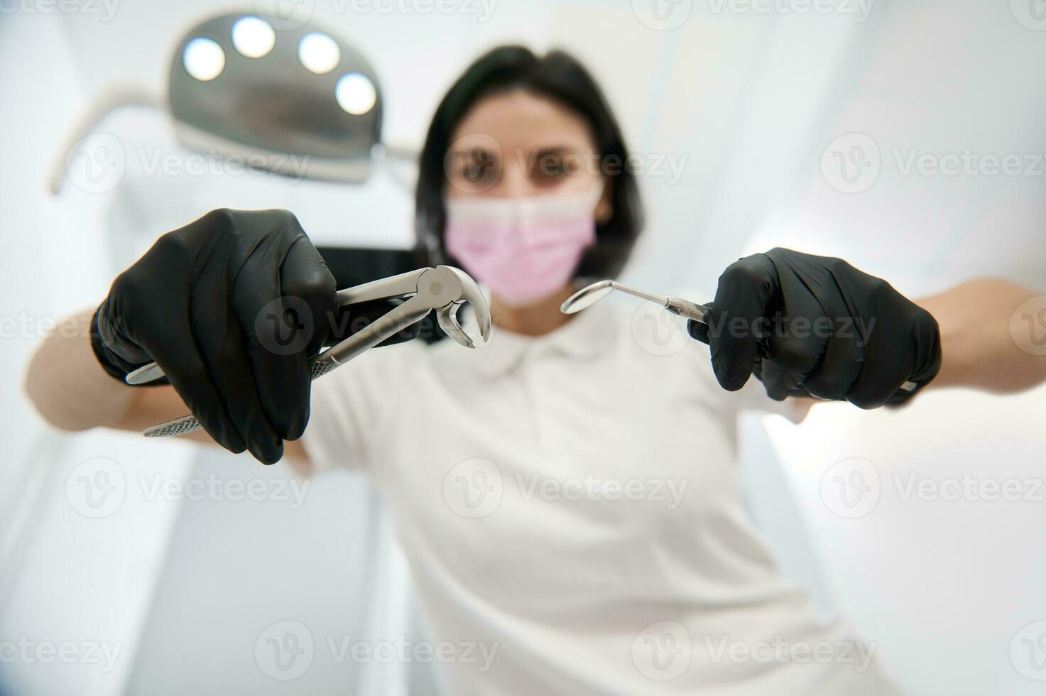 Focus on stainless steel dental instruments, extraction forceps and dental mirror in the hands of blurred dentist in surgical gloves and medical mask leaning towards the patient for an examination photo