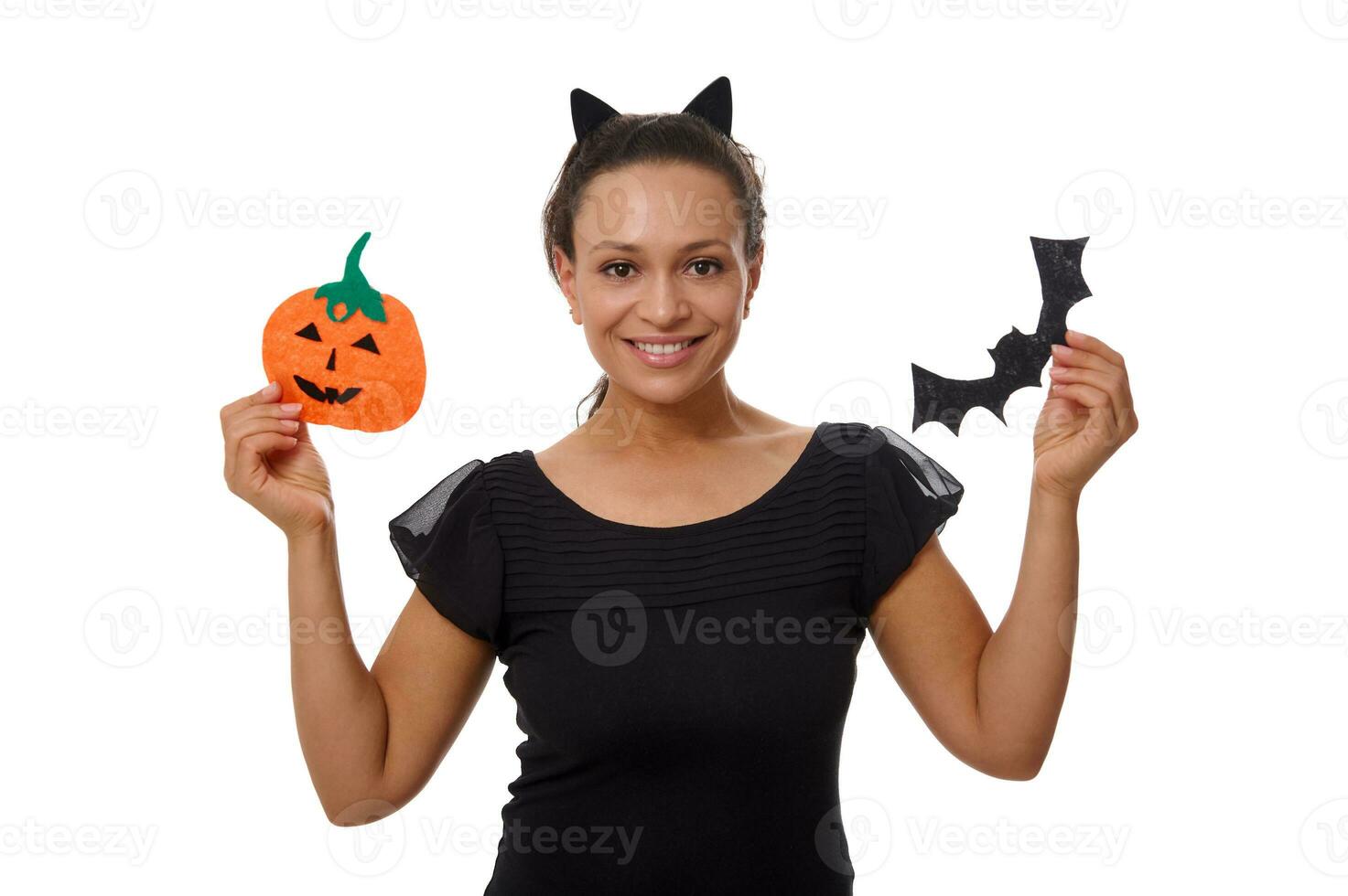 Beautiful woman in cat ears hoop and smiling with cute toothy smile poses against white background with cut felt handmade pumpkin and bat, celebrating Halloween traditional event. Copy space for ad photo