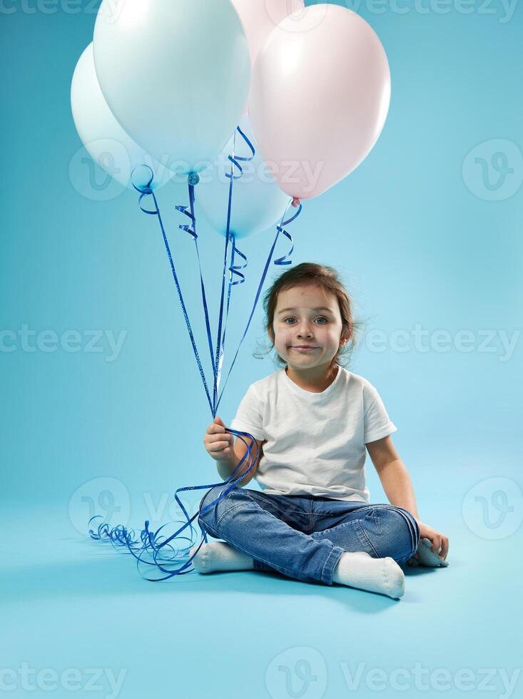 Cute little girl sitting on blue background with soft shadow and posing to camera with balloons in the hand. Childhood and child protection day concept. Copy space photo