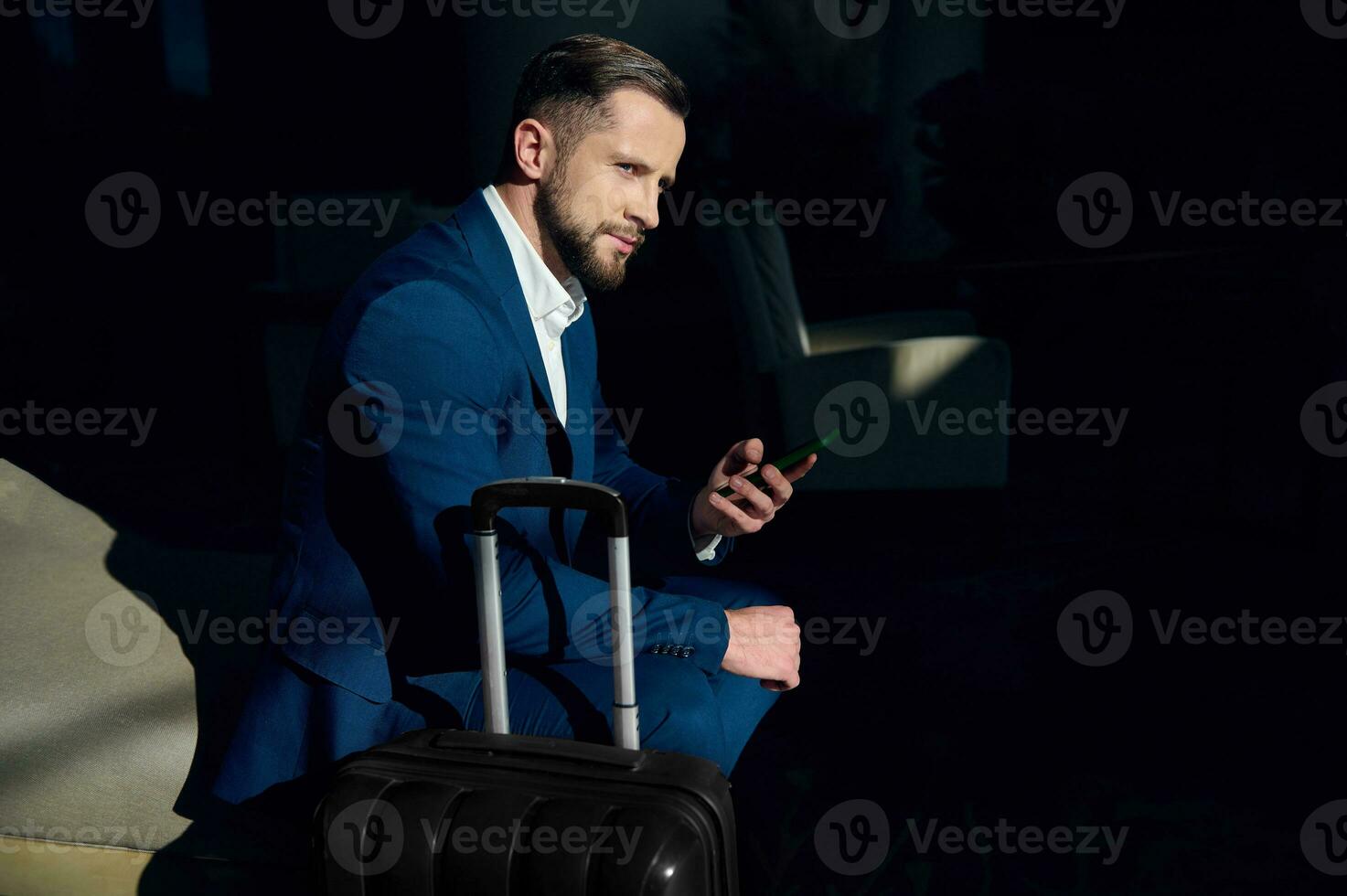 atractivo hermoso hombre, joven exitoso empresario, inversión consultor en un negocio traje, se sienta en un silla siguiente a su equipaje, usos un teléfono inteligente en el hotel vestíbulo durante un negocio viaje foto