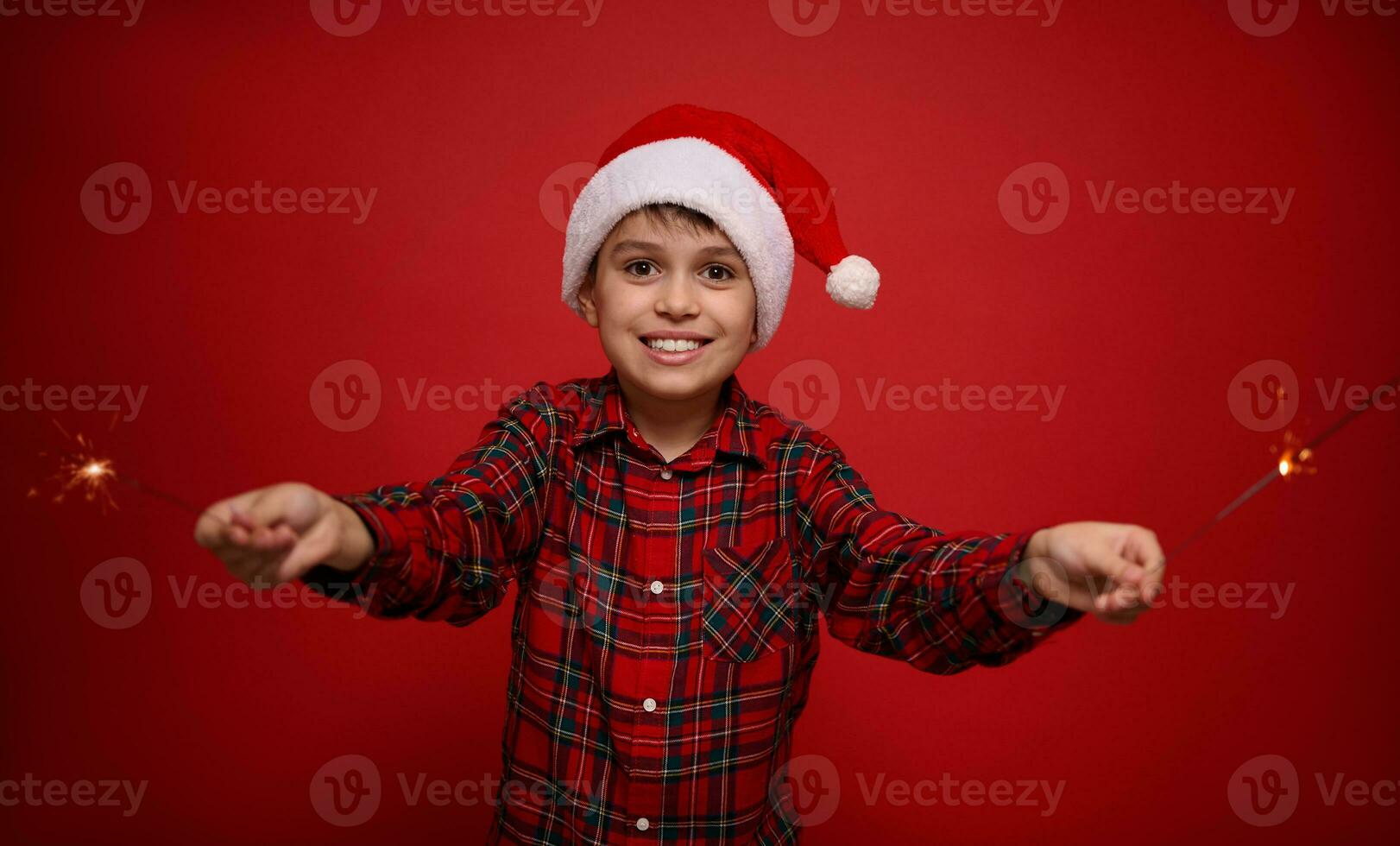 Handsome adorable preteen boy, beautiful child in Santa Claus hat and checkered shirt enjoys the Christmas party, plays with Bengal lights, sparklers, isolated o red background with copy space for ad photo