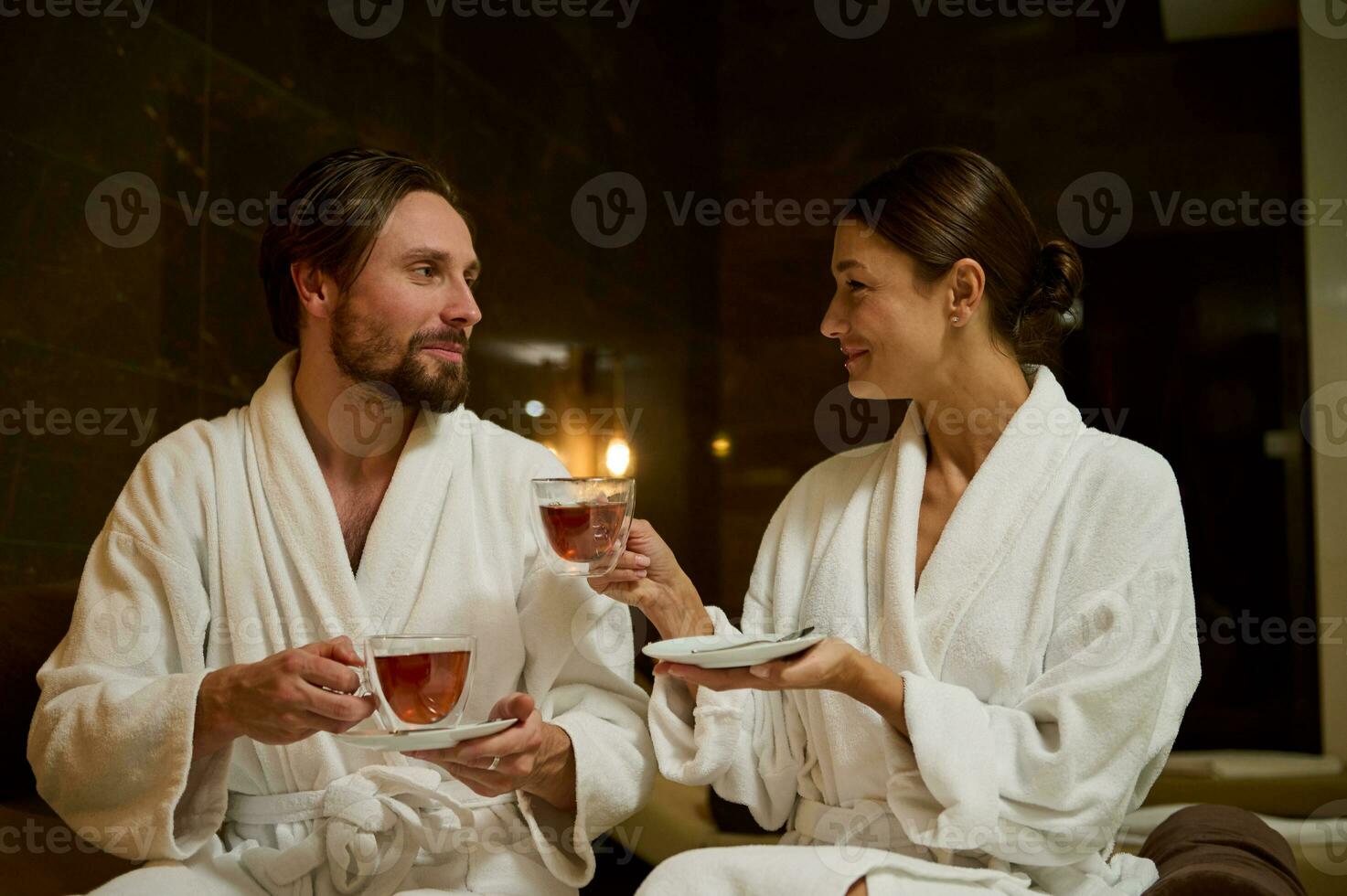 Loving couple in white bathrobes sitting close to each other on a lounge zone by the sea, drinking healthy herbal tea with from cups. Newlyweds on their honeymoon enjoying wellness spa procedures photo