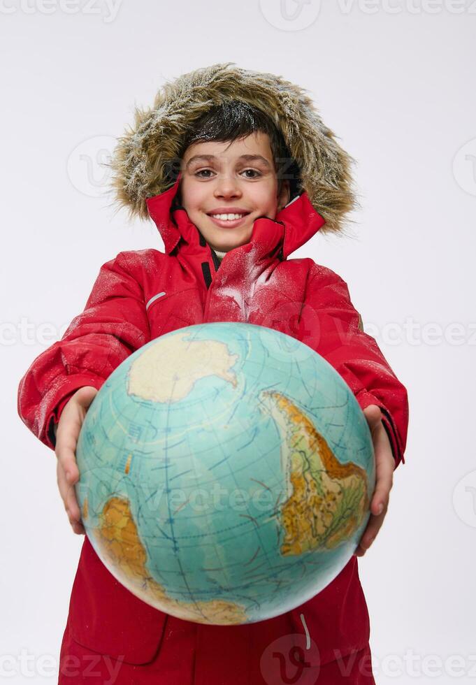 Cheerful European boy in bright red snowy down jacket with hood, holding huge globe in his hands, smiles looking at camera, isolated on white background. Tourism, geography, planet exploration concept photo