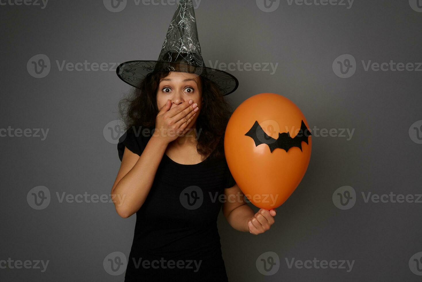 Scared woman dressed in Halloween witch costume, covering her mouth in fear and horror, looking at camera, posing with colorful balloons with felt-cut black bat against gray background with copy space photo