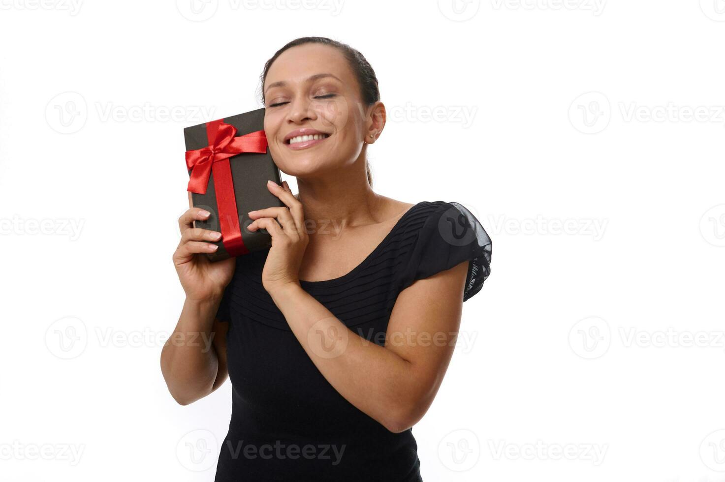 atractivo mujer con cerrado ojos vestido en negro poses a cámara en contra blanco fondo, con negro regalo caja cerca su rostro, sonrisas con dientes sonrisa, disfrutando presente. negro viernes, cumpleaños concepto foto