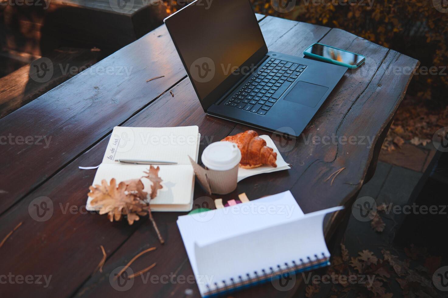 Soft focus on a laptop with copy space on blank black monitor screen near smartphone and out of focus croissant with hot coffee in takeaway paper cup and open copybook and notepad on a wooden table photo