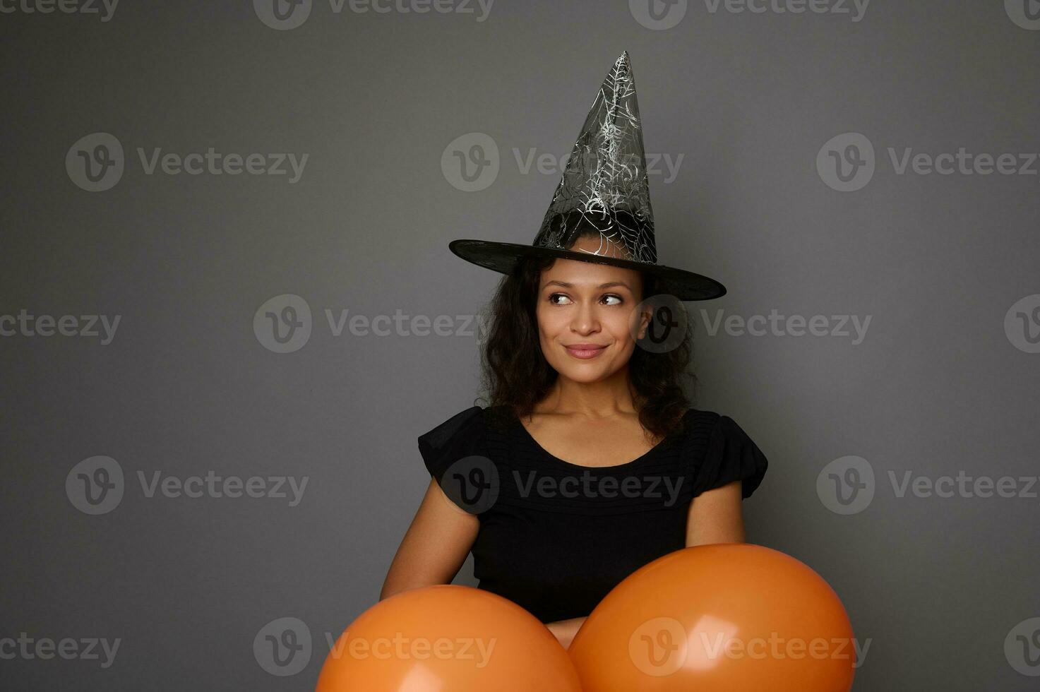 Cute smiling charming Latin American woman dressed in black and witch hat peeks through the orange air balloons, looks to the side to a copy space for ad on gray background. Happy Halloween concept photo