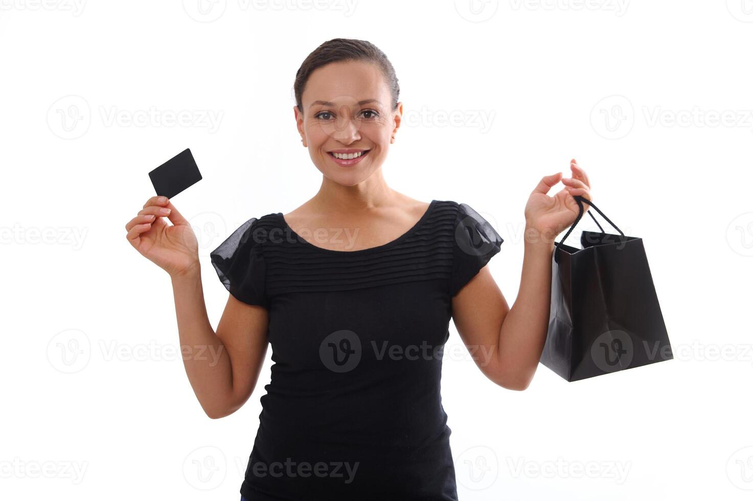 Isolated portrait on white background with copy space of attractive smiling woman dressed in black and holding a discount credit card and a shopping bag, looking at camera. Black Friday concept photo