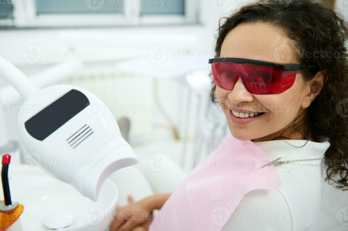 bonito mujer vistiendo uv protector gafas de protección sonrisas con hermosa con dientes sonrisa sentado en dentistas silla, mirando a cámara después dientes blanqueo procedimiento con especial uv ligero lámpara en un dental clínica foto