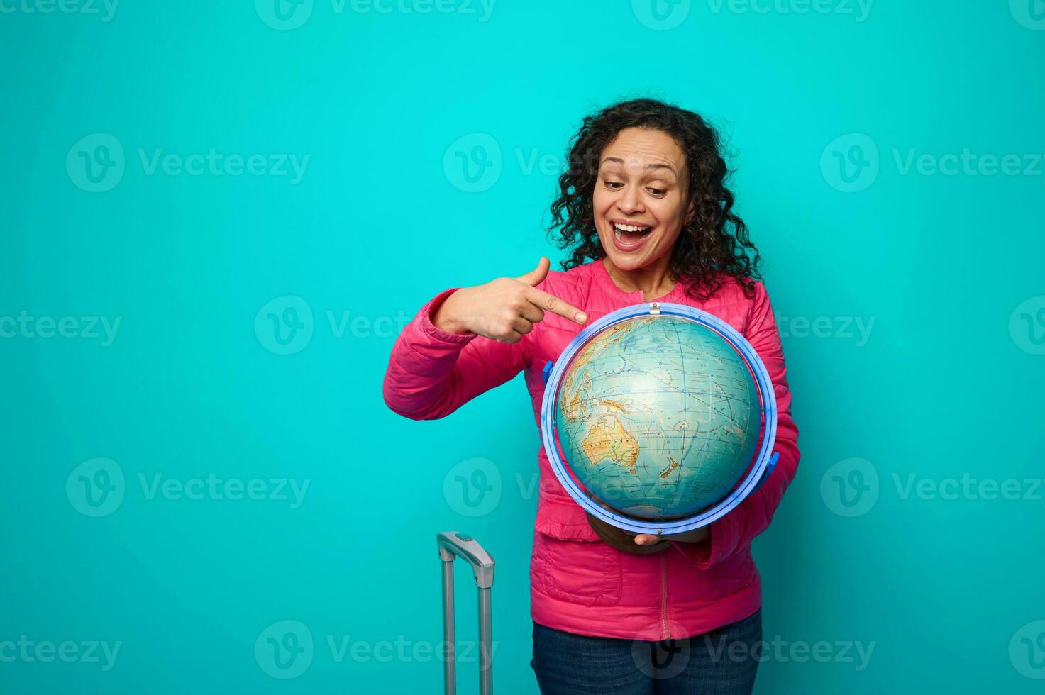 Cheerful delightful woman in pink jacket points with finger to travel destination on the map of a globe in her hands. Concept of tourism, travel around the world with copy ad space on blue background photo