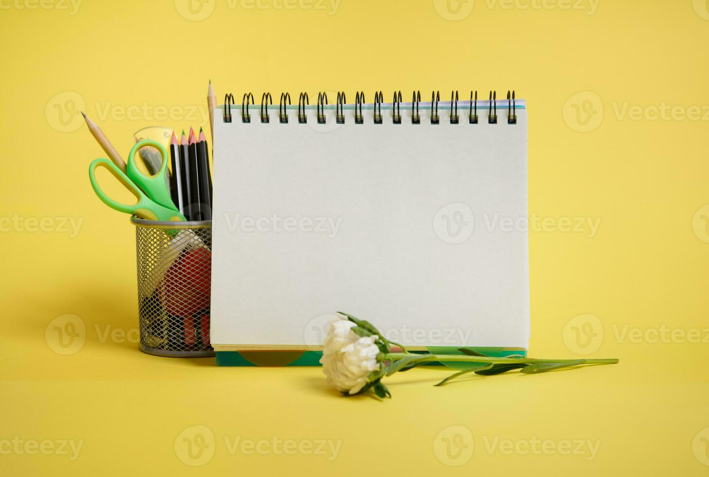 Close-up of a metal bucket with stationery. Holder with colored school supplies, empty blank white paper sheet of an organizer and aster flower lying down on yellow background with copy space photo