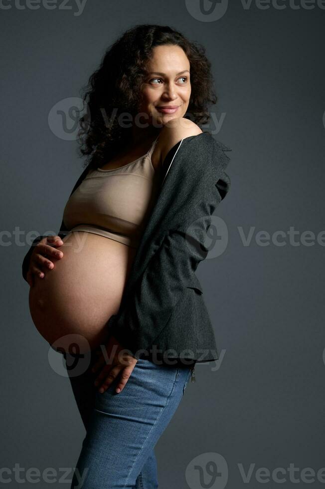 retrato de un elegante elegante embarazada mujer poses desnudo barriga, sonrisas mirando aparte, aislado en gris estudio antecedentes. foto